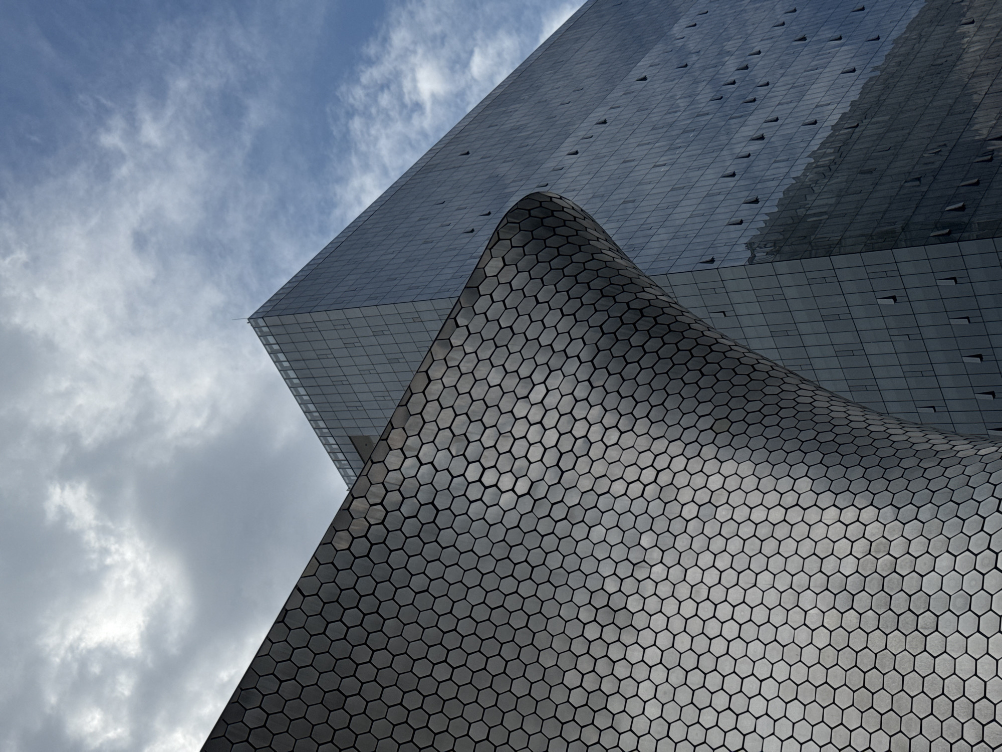 The image captures a low-angle view of two striking architectural structures in Mexico City, Mexico, set against a partially cloudy sky. The foreground features a section of a building with a distinctive curved surface covered in hexagonal tiles. The color of the building is a muted grey. Behind the curved structure, a portion of a taller, rectangular building with a grid-like facade is visible, suggesting a modern design. The sky is a mix of light blue and grey clouds, providing a contrasting backdrop to the geometric forms of the buildings. The lighting appears diffused, highlighting the textures and patterns of the building materials.