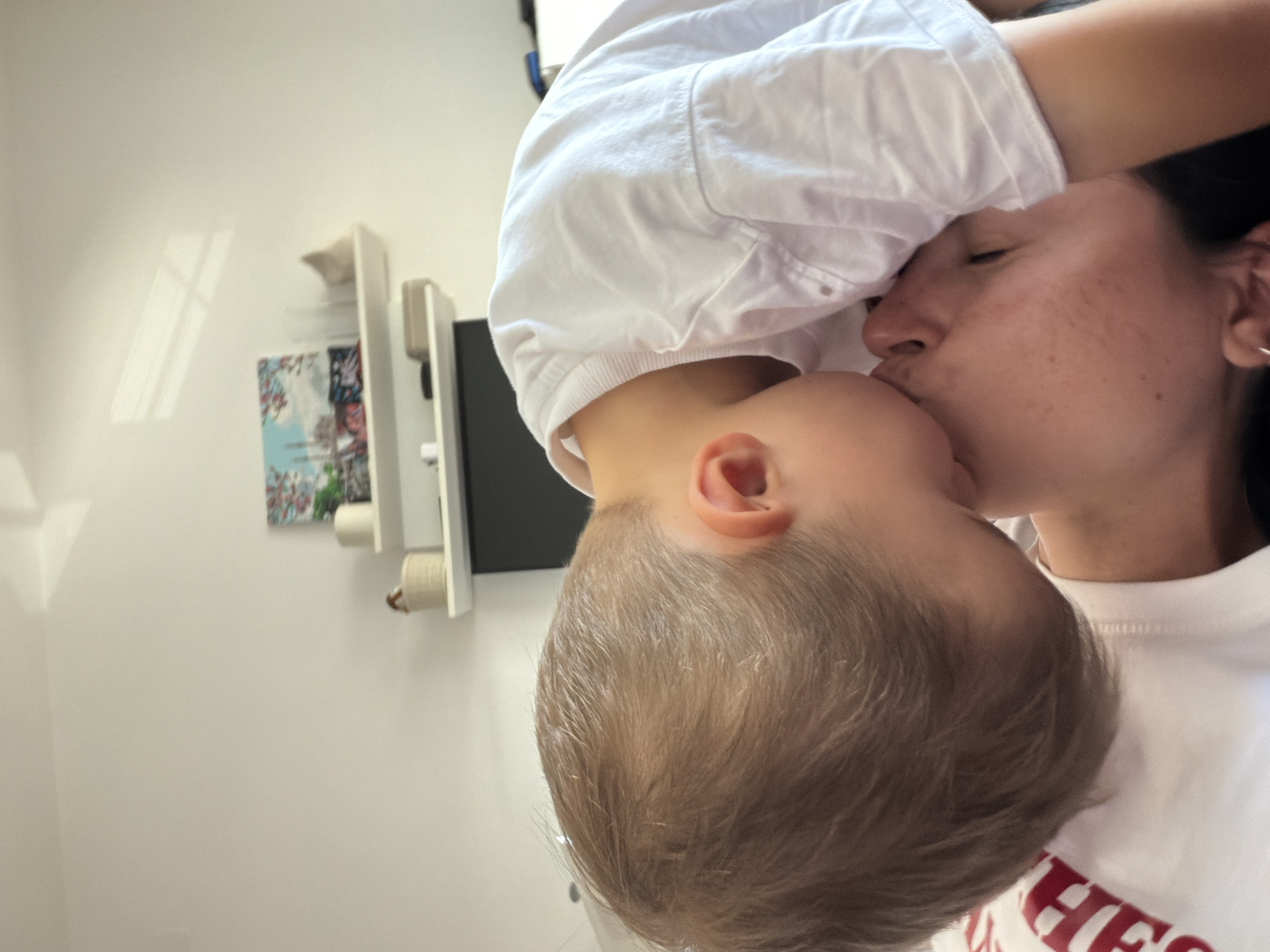 The image captures an intimate indoor scene featuring two individuals: an adult and a young child, likely embracing or sharing a kiss. A fair-skinned child with light brown hair, wearing a white t-shirt, is positioned leaning over the adult, pressing their mouth against the adult's nose and upper cheek area. The adult, also fair-skinned, is lying down with their head slightly tilted to the right, showing a profile view of their face, including an ear with a small silver hoop earring. They are wearing a white garment with partially visible red lettering, possibly an "H."
The background consists of a plain, light-colored wall. On the wall, two white floating shelves are mounted. The upper shelf holds a light-colored cylindrical object and a small woven basket. The lower shelf displays a framed artwork depicting a blue sky, possibly a building or structure, with pink floral embellishments, a clear glass, and a small, light-colored decorative object. Below this shelf, a dark rectangular screen, possibly a television, is partially visible. Strong natural light from an unseen window casts distinct rectangular shadows onto the upper part of the wall. No specific visual cues are present within the image to identify the location as Budva, Montenegro, or any other specific city.