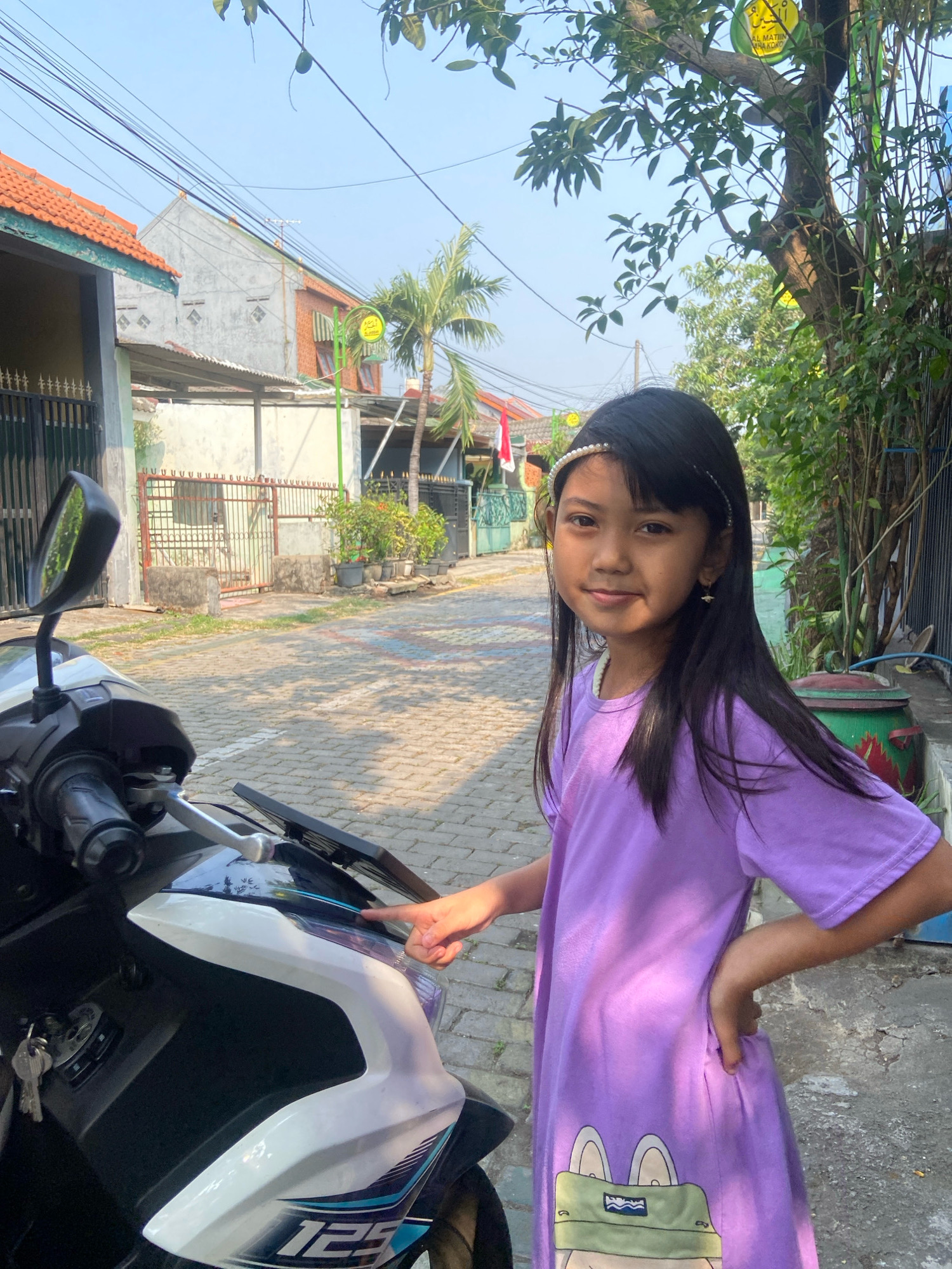 In Gelam, Indonesia, a young girl with long, dark hair and a pearl headband stands next to a scooter on a paved street. She is wearing a purple t-shirt dress with a cartoon frog graphic. Her right hand is on her hip, and her left hand is pointing at the scooter. The background shows houses with orange tile roofs, fences, and lush green trees. The scene is illuminated by bright sunlight, casting shadows on the ground. A round green sign with Arabic writing is visible on a pole behind the girl.