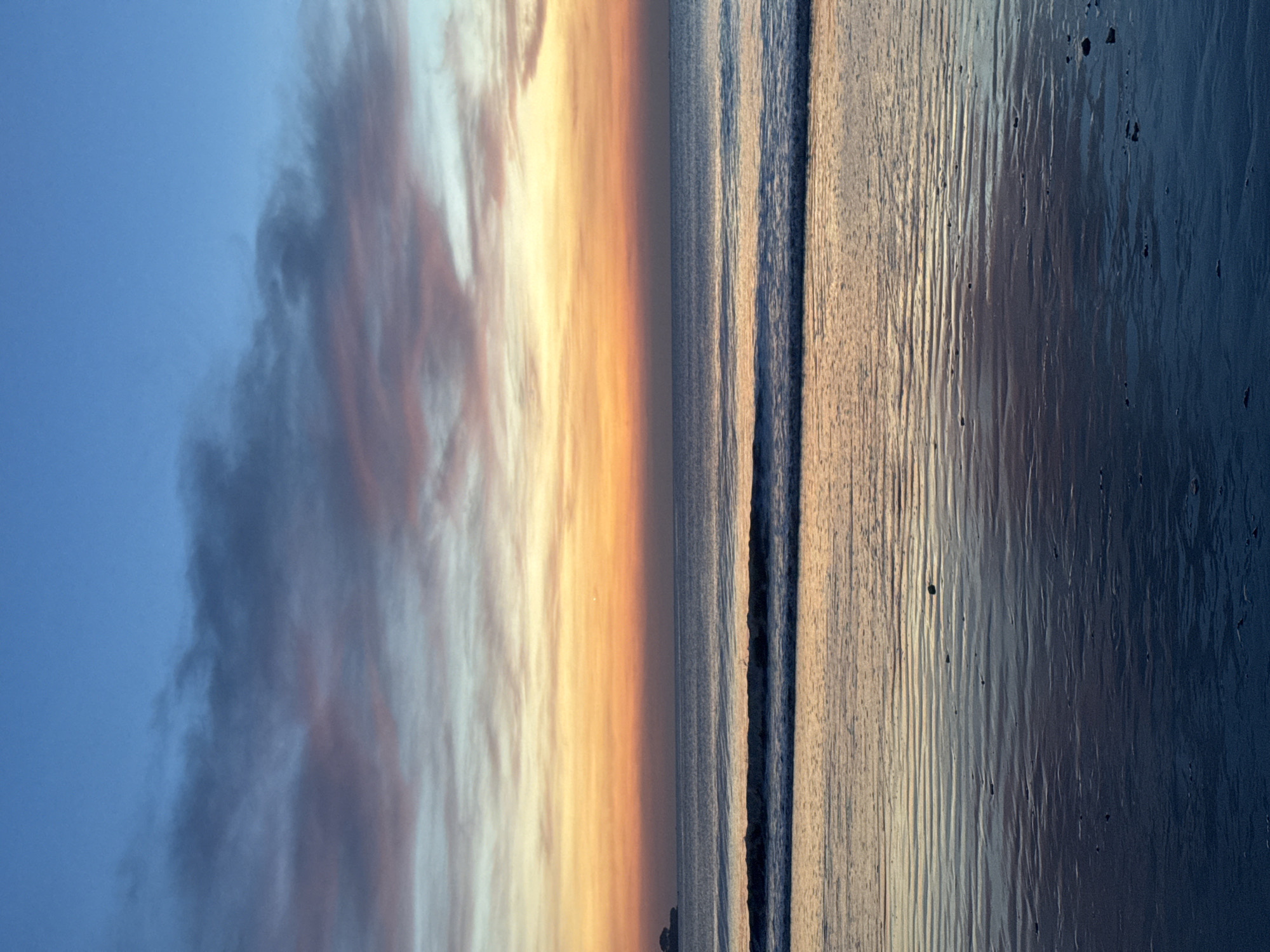 Here is a description of the image:
The image shows a beach scene at dusk. The sky transitions from blue to orange and yellow, indicating either sunrise or sunset. Clouds are present on the left side of the frame. In the center is a strip of ocean with small waves. On the right, the wet sand reflects the sky, creating a mirror effect. The scene is likely in Kuta Selatan, Indonesia, based on the visual characteristics of the beach and lighting conditions. There are no people visible.