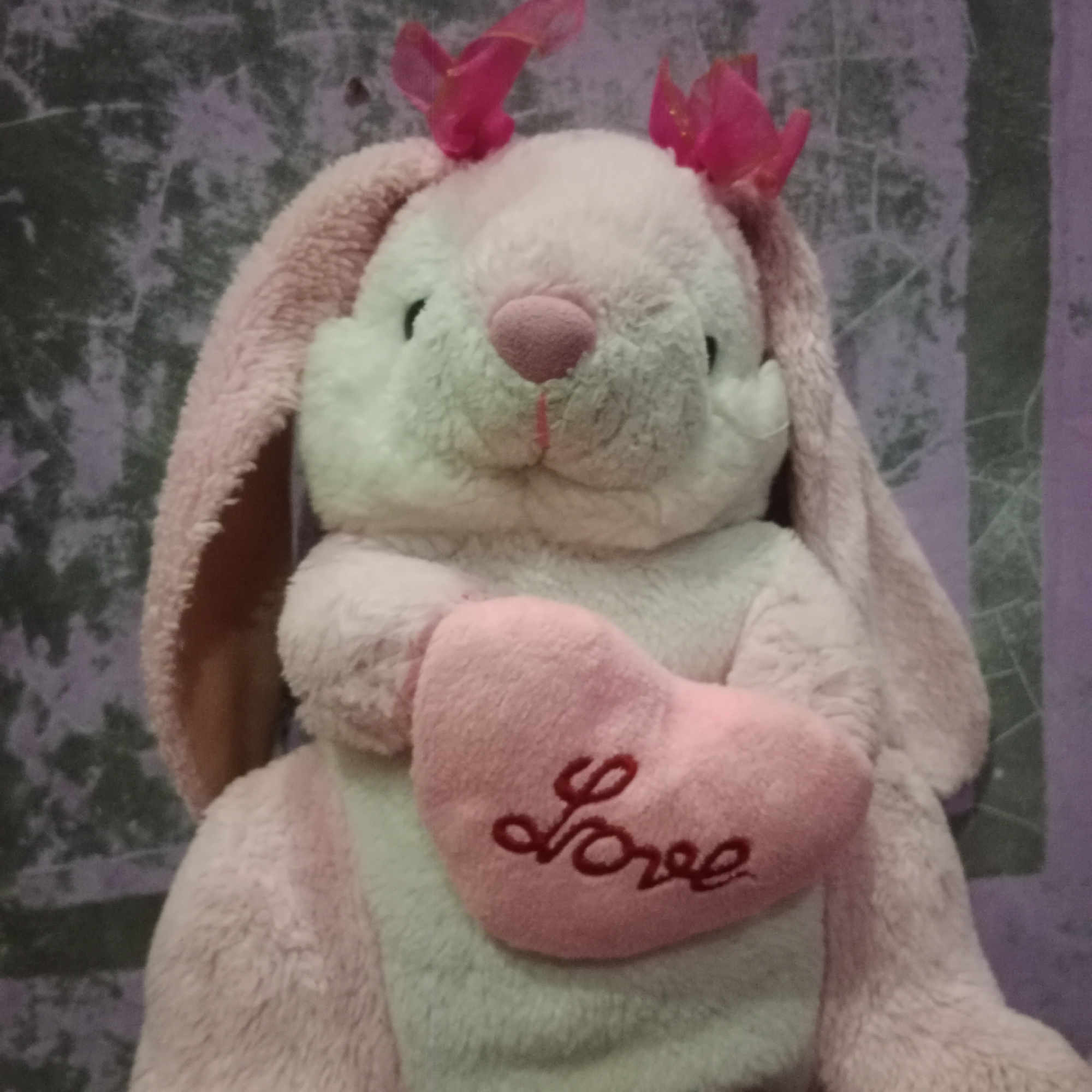 A plush pink bunny with white accents and a pink heart with the word "love" embroidered in red sits against a textured purple background. The bunny has pink ribbon bows on its head and long, floppy ears. The heart is held in the bunny's paws. The background is a mottled pattern of dark and light purple. The image is a close-up, focusing on the bunny. There is no indication of the city of Tegowanu Kulon, Indonesia in the image.