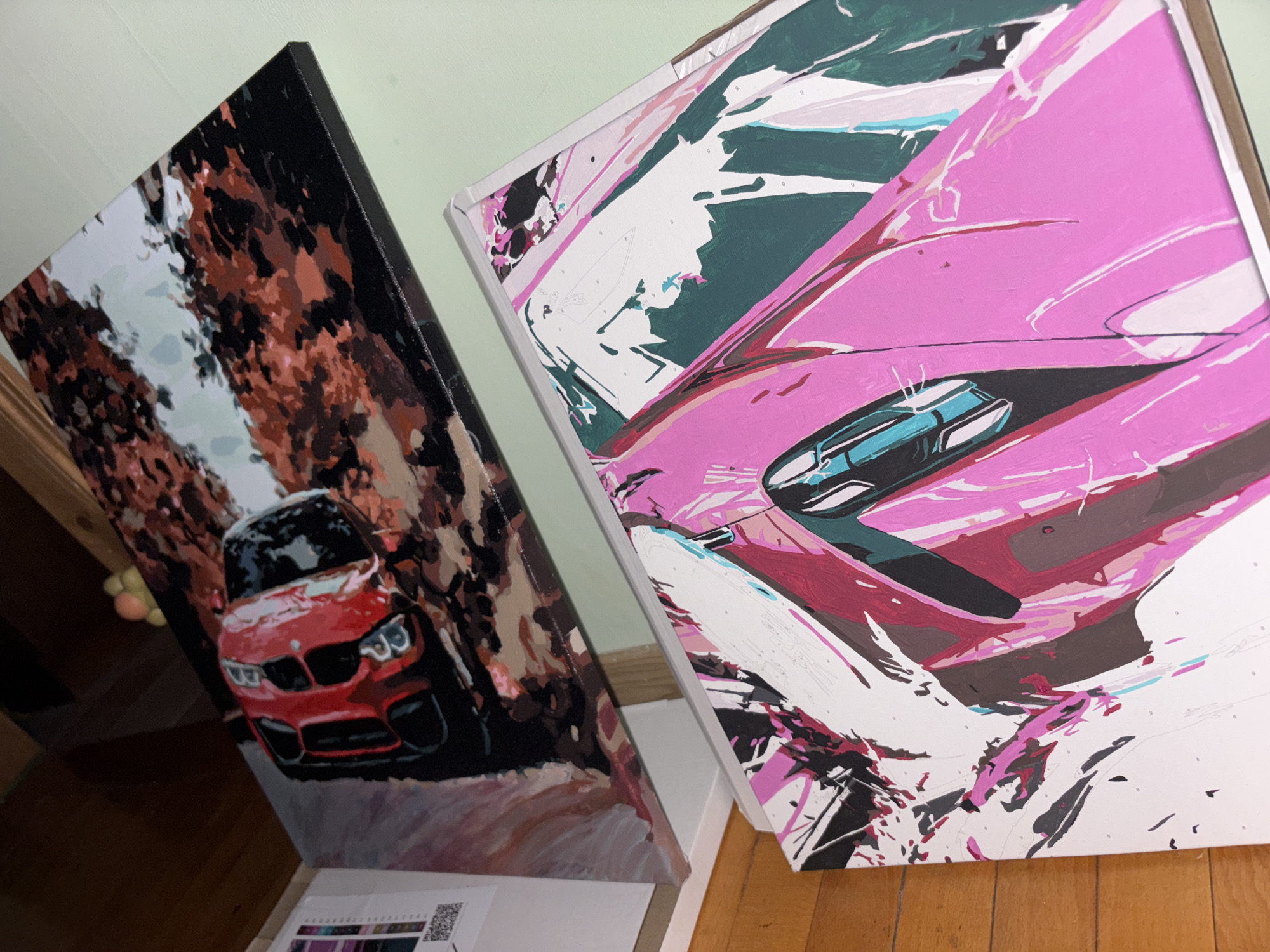 Two paintings are propped up against a wall, one depicting a red car driving on a road surrounded by brown trees, and the other showing a pink car with abstract brushstrokes in shades of pink, white, black, and green. The paintings are resting on a white surface, and a wooden floor is visible beneath them. A color palette and a QR code are visible in the lower center of the image. The location is not specified in the image and therefore cannot be confirmed as Vsevolozhsk, Russia.