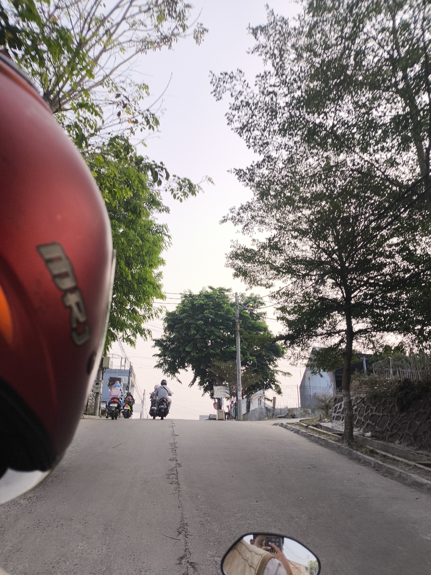 The image depicts a street scene in Cilegon, Indonesia, taken from the perspective of someone riding a motorcycle. In the foreground, a red motorcycle helmet is visible on the left side. The road ahead is paved with asphalt and shows signs of wear, including cracks and patches. Several motorcycles are traveling up the incline of the road. On the right side of the road, a stone wall and vegetation are present. Trees line both sides of the road, with the sky visible in the background. The lighting suggests it is either early morning or late afternoon. In the motorcycle's rearview mirror, a reflection of a person taking a photograph with a smartphone is visible.