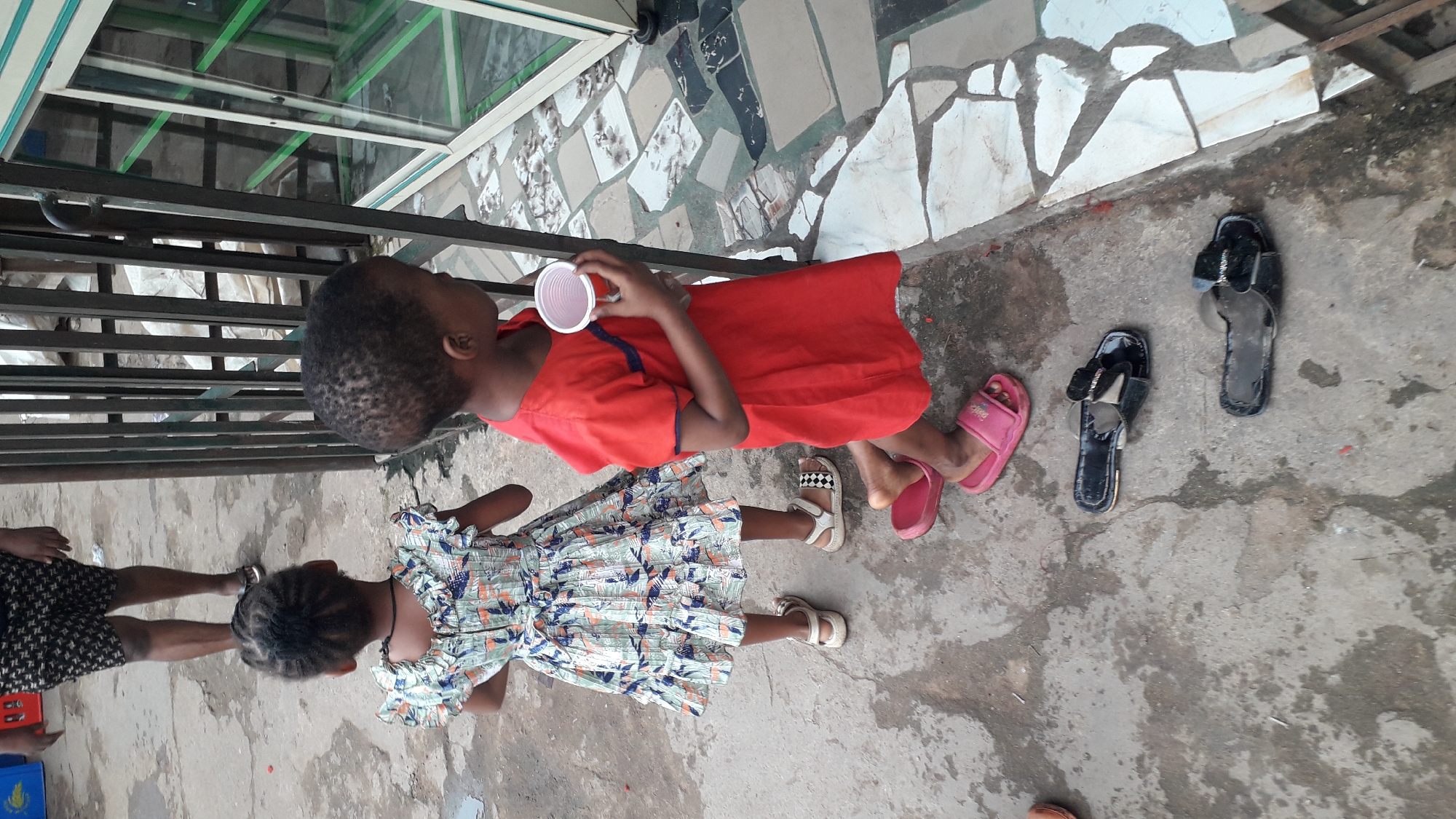 Two children, one in a red dress and the other in a patterned dress, stand on a concrete surface. The child in red holds a white plastic cup. Nearby are pairs of sandals and slippers. A tiled area with irregular patterns is visible in the background. A metal railing and a glass-enclosed structure are also present. The scene is located in Onitsha, Nigeria.