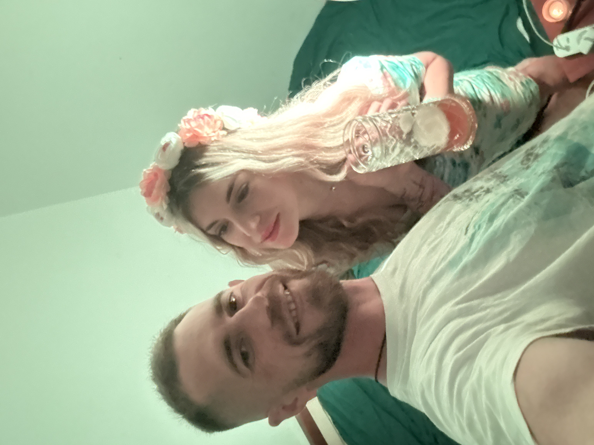A close-up indoor selfie shows a light-skinned man and woman. The man is in the foreground, wearing a white t-shirt and sporting a beard and mustache. The woman, positioned behind him, wears a floral crown on her light blond hair and holds a glass with an amber-colored beverage and ice. The background includes a green blanket. The location is unknown.