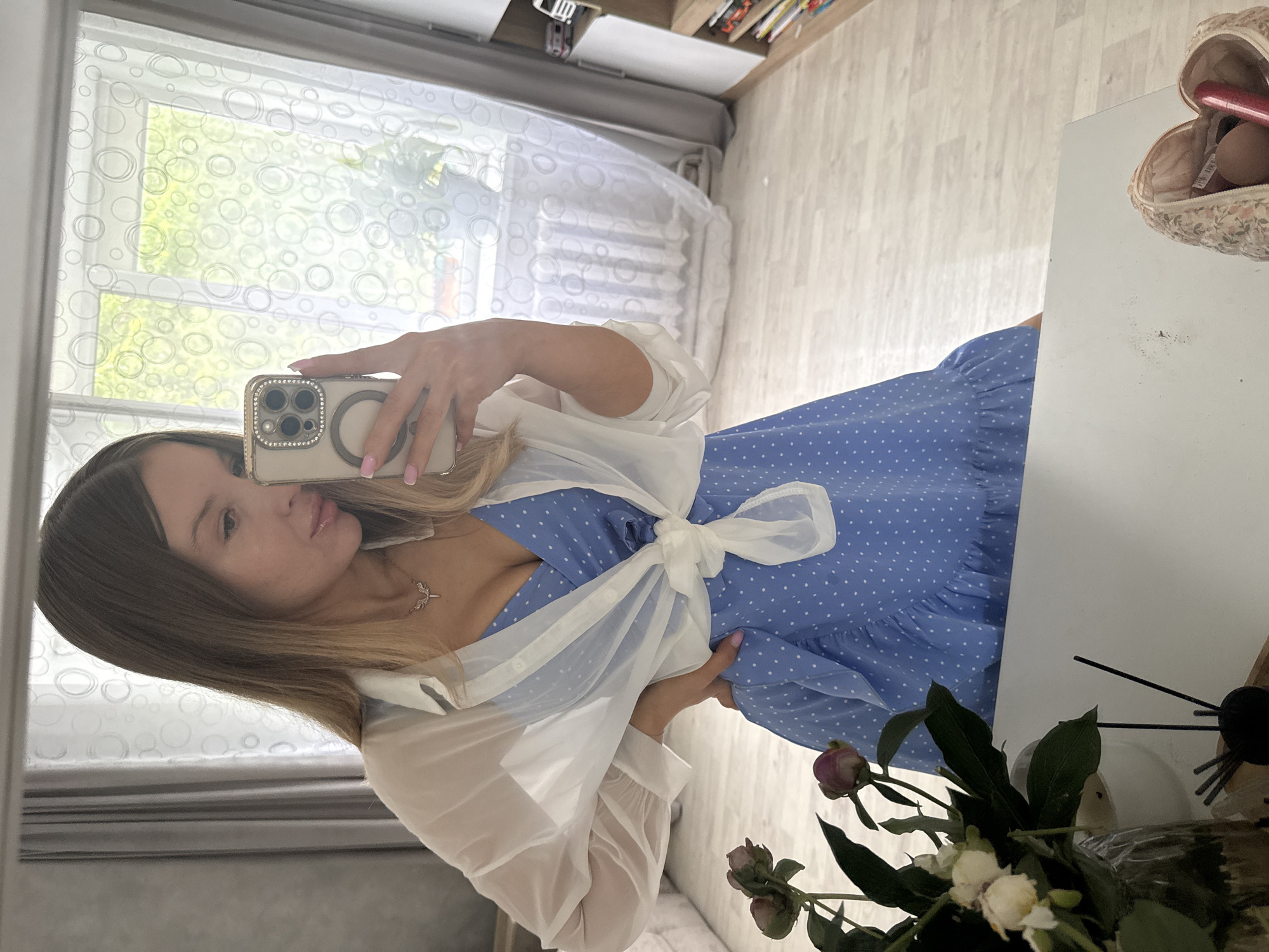 A young woman takes a mirror selfie in a room with light-colored walls and flooring. She is wearing a blue polka-dot dress with ruffled hem and a sheer white open shirt tied at the waist. A delicate necklace with a winged pendant is visible on her chest. Her hair is light brown and styled long. To the left of the frame, a bouquet of peonies in a glass vase is visible, along with a black diffuser. In the foreground, a part of a table with a quilted makeup bag containing cosmetics is visible. The background shows a window with patterned curtains and a radiator, and a bookshelf with toys and books. The overall impression is a domestic indoor setting, possibly a bedroom or living area. There is no indication of the specific city of Октябрьское, Russia.