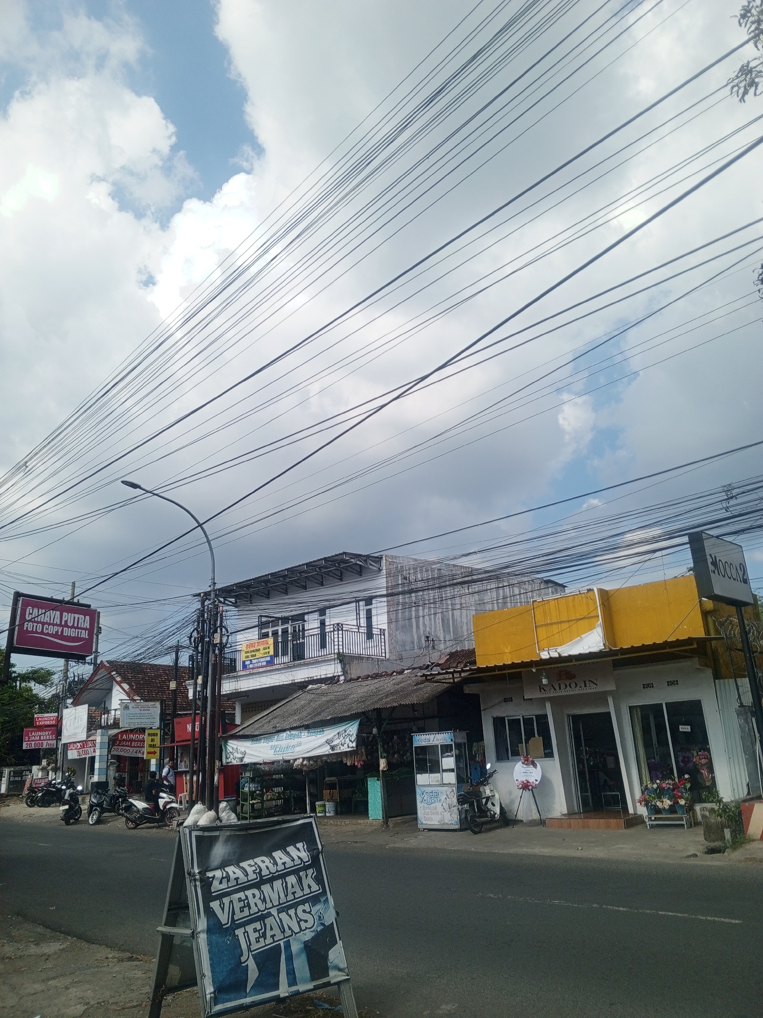 The image depicts a street scene in Sumbersari, Indonesia, dominated by a dense network of overhead power lines against a partly cloudy sky. The foreground shows a paved road with a white dashed line. On the right side of the road, a white building with a bright yellow awning is visible, featuring glass doors and windows. A sign above the doors reads "KADO.IN". To the left of this building, a blue and white A-frame sign stands on the sidewalk with the text "ZAFRAN VERMAK JEANS" printed in large, bold letters.
Further down the street on the left, a series of businesses are lined up. One building has a prominent red and white sign that says "CAHAYA PUTRA FOTO COPY DIGITAL". Other signs, partially obscured, advertise laundry services and other shops. Several motorcycles are parked along the sidewalk in front of these businesses. A street lamp is visible on the left side of the image. The buildings appear to be of moderate height, with some having balconies. The overall impression is a commercial street in a town or city.