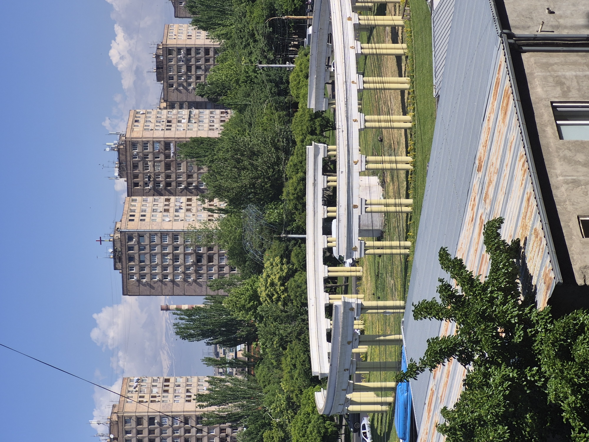 An urban landscape captured during a sunny day in Yerevan, Armenia. The scene is dominated by multiple multi-story apartment buildings with numerous windows and balconies, characteristic of Soviet-era architecture. These buildings, ranging in color from light beige to darker grey, are visible in the background and mid-ground, interspersed with lush green trees. One building in the distance features a small cross on its rooftop. Further in the background, a tall, slender industrial chimney can be seen.
In the mid-ground, a prominent white elevated structure with yellow cylindrical supports runs horizontally across the frame. This structure has a distinct modern, curved design, possibly an elevated walkway or a decorative architectural element. Below and around this structure are grassy areas and more green trees, some showing lighter green foliage indicating new growth. Utility poles and wires are also present, crisscrossing the scene.
The foreground is occupied by the rooftops of buildings. To the right, a dark concrete roof section is visible with a single window. Adjacent to it is a light grey corrugated metal roof, showing patches of rust, which adds texture to the image. A dense, green tree partially obscures this roof in the lower right corner. A white car and a blue container are partially visible near the base of the elevated structure on the left side of the foreground.
The sky is a clear, bright blue with scattered white cumulus clouds. Strong sunlight indicates daytime, casting visible shadows and illuminating the scene vibrantly. There is no discernible text in the image. The overall impression is one of a bustling city with a mix of residential structures and green spaces under a wide, open sky.