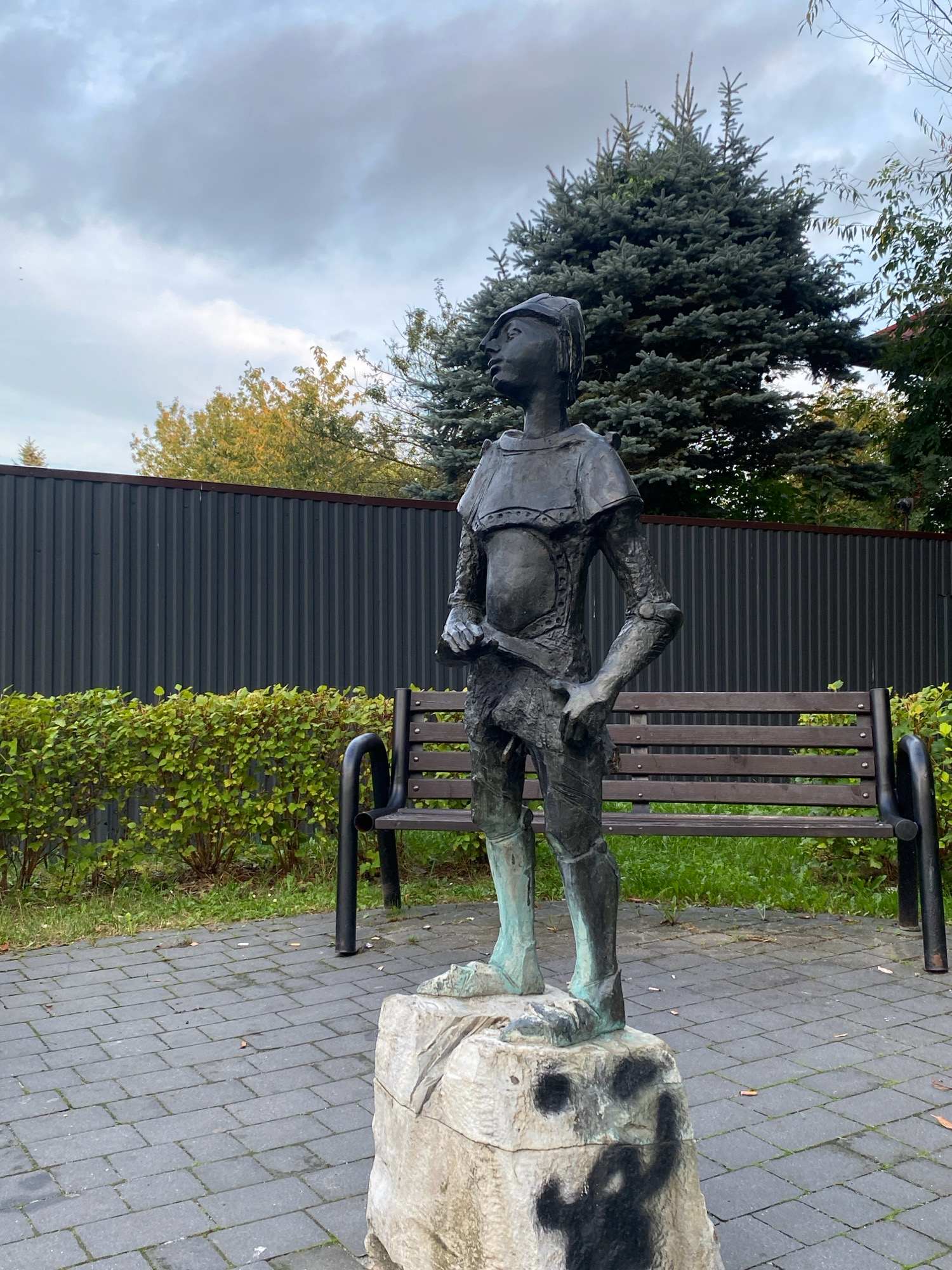 The image shows a bronze statue of a young boy in medieval-style attire standing on a rough, light-colored stone base. The statue is positioned outdoors on a paved area made of dark grey rectangular bricks. The boy wears a helmet, a tunic with armor-like detailing, and what appear to be greaves on his lower legs. He is looking upwards and to his left, with his hands resting at his sides, one holding a long, slender object.
Behind the statue, a dark grey, corrugated metal fence runs horizontally across the frame. In front of the fence and behind the statue, there is a neatly trimmed green hedge. A dark wooden park bench with black metal supports is situated behind the statue, partially obscured by the hedge. Behind the bench and hedge, a large evergreen tree with dense, dark green foliage dominates the background, with a few deciduous trees with yellowing leaves visible to its left. The sky above is cloudy with a mix of grey and white.
The statue is located in Nowy Sącz, Poland. This specific statue is a memorial to Janosik, a legendary folk hero and outlaw of the Slovak and Polish mountains.