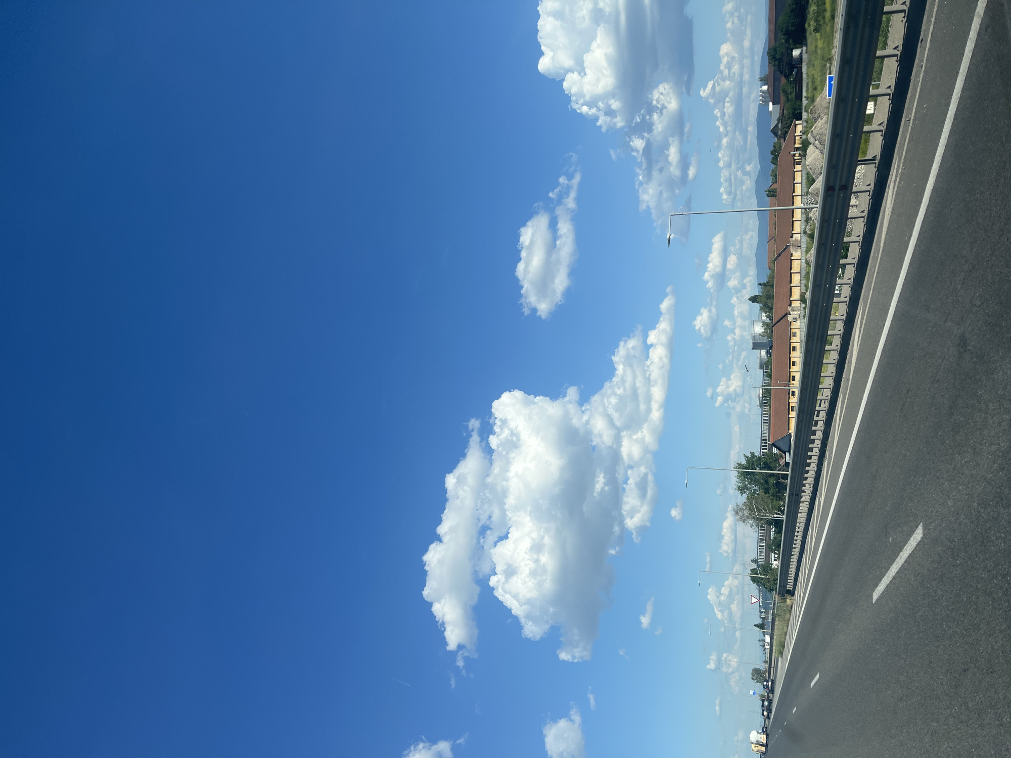 The image depicts a view from a moving vehicle on a highway, looking towards the sky and the right side of the road. The sky is a vibrant, clear blue, with scattered fluffy white clouds. The highway itself is paved with asphalt and has white lane markings. A metal guardrail separates the road from the adjacent area.
On the right side of the guardrail, there are some buildings with orange-brown roofs and yellow facades, suggesting an urban or suburban setting. Trees and vegetation are visible, adding to the scenery. Several streetlights are positioned along the highway. In the distance, mountains can be seen. The image appears to be taken during the daytime, likely late morning or early afternoon, given the bright sunlight.
The perspective suggests the photo was taken from the passenger seat or driver's side window of a car. There are no people clearly visible in the image, and the focus is on the landscape and infrastructure. The overall mood is peaceful and serene, characteristic of a scenic drive.
There is some text visible on a blue road sign.