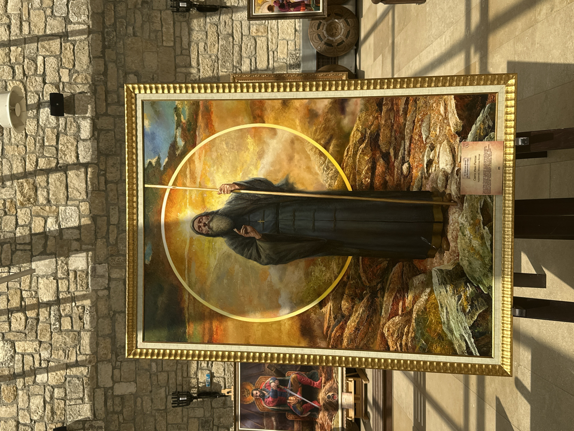 The image shows a large framed painting displayed on an easel in a room with a stone wall. The painting depicts an elderly man with a long beard, wearing dark robes and holding a staff. He is standing in front of a golden circle and a dramatic sky. A small plaque is attached to the bottom of the frame, which appears to be an informational label for the artwork. To the left of the main painting, another framed artwork is visible, showing a man in a red and blue outfit seated on a throne, with a sword in his lap. In the background, other artworks and decorative items are visible, as well as a sign with the word "EXIT". The overall setting suggests an art exhibition or a museum. The question specifies the location as "Neofit Rilski, Bulgaria." However, the image itself does not contain any explicit visual cues to confirm this specific location beyond the presence of the plaque which may contain relevant information.