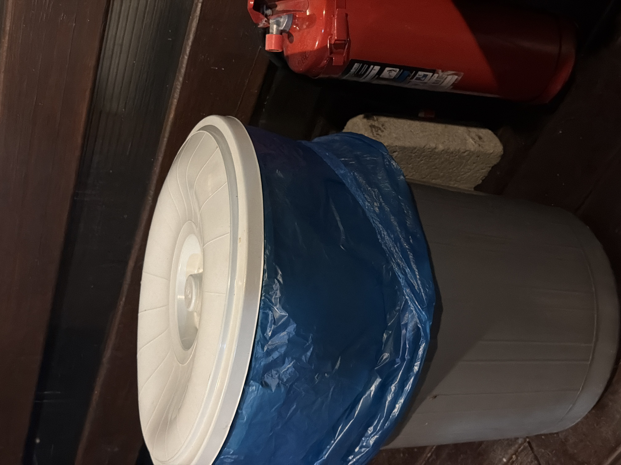 A grey trash can with a white lid and a blue trash bag is positioned on a wooden surface next to a red fire extinguisher. A light-colored concrete block is partially visible behind the trash can. The background consists of dark wooden slats. The scene is dimly lit, suggesting it might be outdoors or in a poorly lit indoor area. The overall impression is of a utilitarian setting. The image does not contain any identifiable landmarks or clues to definitively place it in Sochi, Russia.