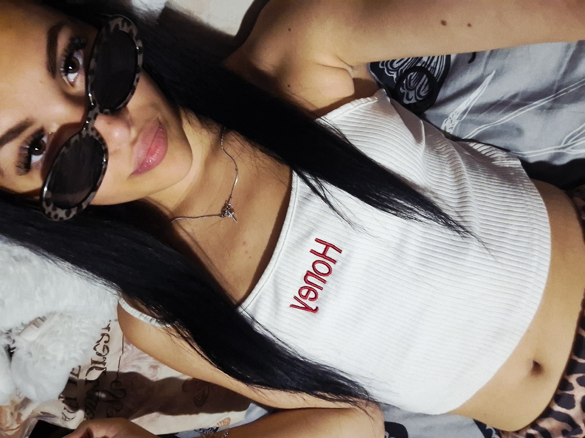 A young woman is captured in a close-up, high-angle selfie, lying down indoors, likely in a bedroom setting in Krapets, Bulgaria. She has long, straight dark hair. Her eyes are visible above leopard print-framed sunglasses, which rest on her nose. She wears a white, ribbed crop top that exposes her midriff, with red embroidered text on the chest that reads "VƏNOH" (HONEY, reversed). A thin silver necklace with a small pendant adorns her neck. The bedding beneath her features various patterns, including a grey sheet with a black and white abstract design and a brownish/cream sheet with a white, fluffy texture. A leopard print pattern, possibly on her lower garment or additional bedding, is visible on the right. Her left hand is seen at the bottom left, holding what appears to be a phone. Her expression is calm with a subtle smile. The lighting suggests an indoor environment, possibly illuminated by artificial light.