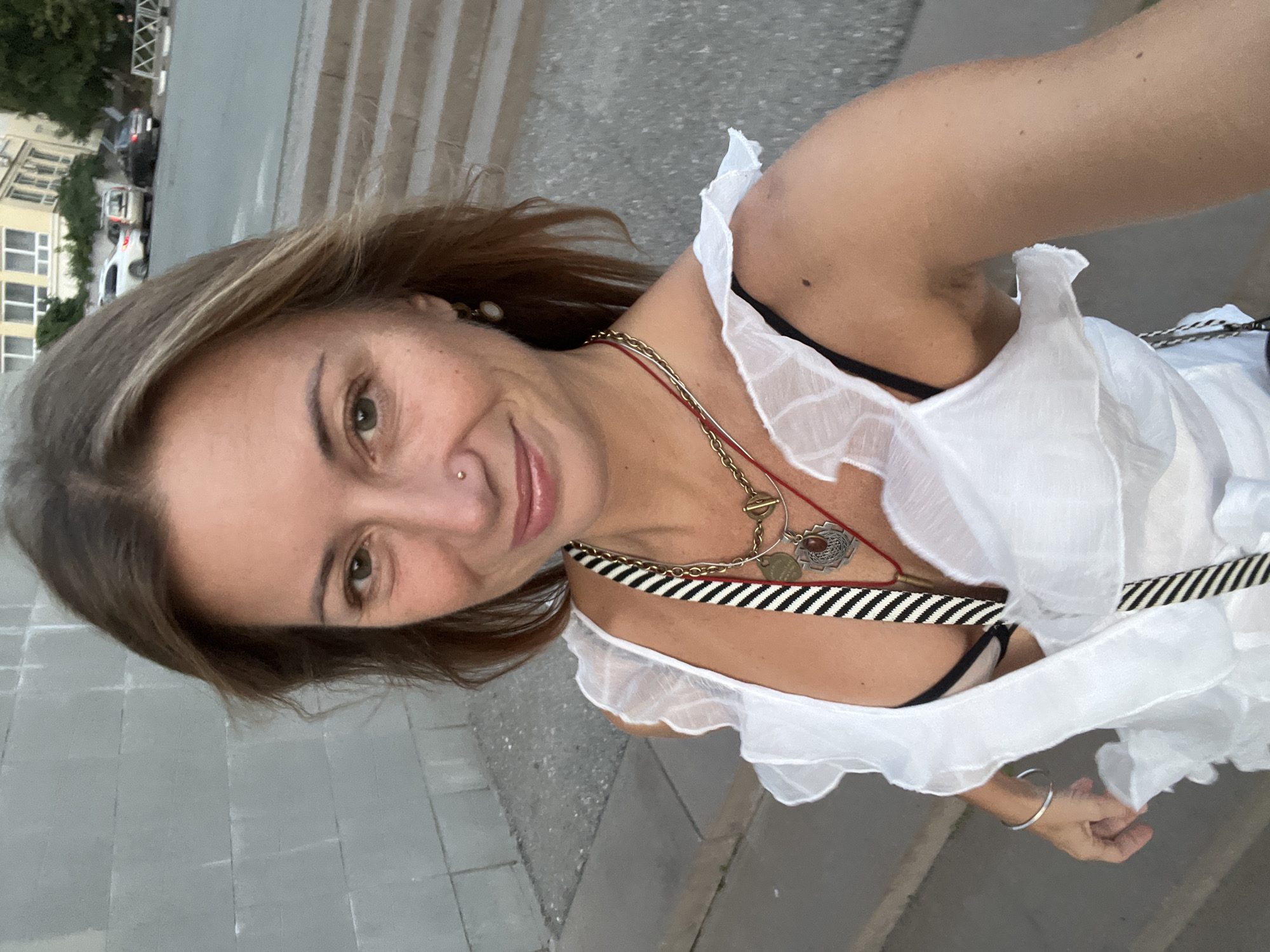 A woman with brown hair and green eyes is taking a selfie outdoors. She is wearing a white dress with ruffled sleeves and a black bra. She has several necklaces on, including a gold chain with a pendant. She is also wearing a nose ring and an earring. The background shows a concrete wall, stairs, and a street with cars. The location is Samara, Russia.