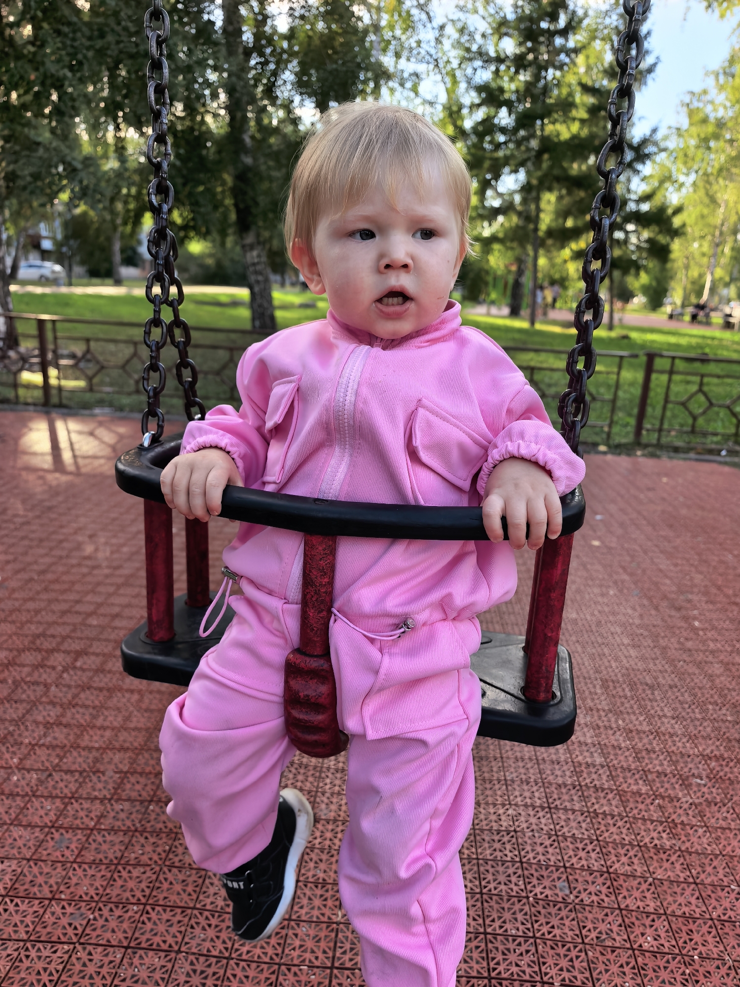 A young child with blonde hair, wearing a pink tracksuit and black sneakers, is sitting on a swing set. The swing has black and red components. The ground is a red, textured surface. In the background, there are trees, a fence, and a street with a car, suggesting an outdoor park or playground setting within the city of Sterlitamak, Russia. The lighting indicates it is daytime.