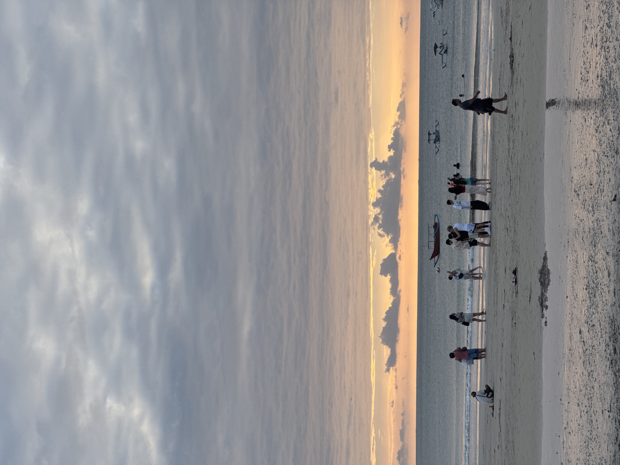 The image depicts a beach scene in Kuta Selatan, Indonesia, during what appears to be sunset. The sky is filled with layered grey and pink clouds, with a warm orange glow near the horizon. The ocean is calm, with small waves lapping the shore. Several boats, including some traditional outrigger boats, are visible on the water.
On the sandy beach, a group of people are scattered. Some are standing, some are walking, and one person is sitting down. There is a group of individuals gathered near the water's edge, some of whom appear to be taking photos. Another person is walking along the shoreline in the foreground, and a lone individual is seated on the sand further back. The sand itself shows signs of wetness and reflection from the water. The overall atmosphere suggests a relaxed and tranquil evening at the beach.