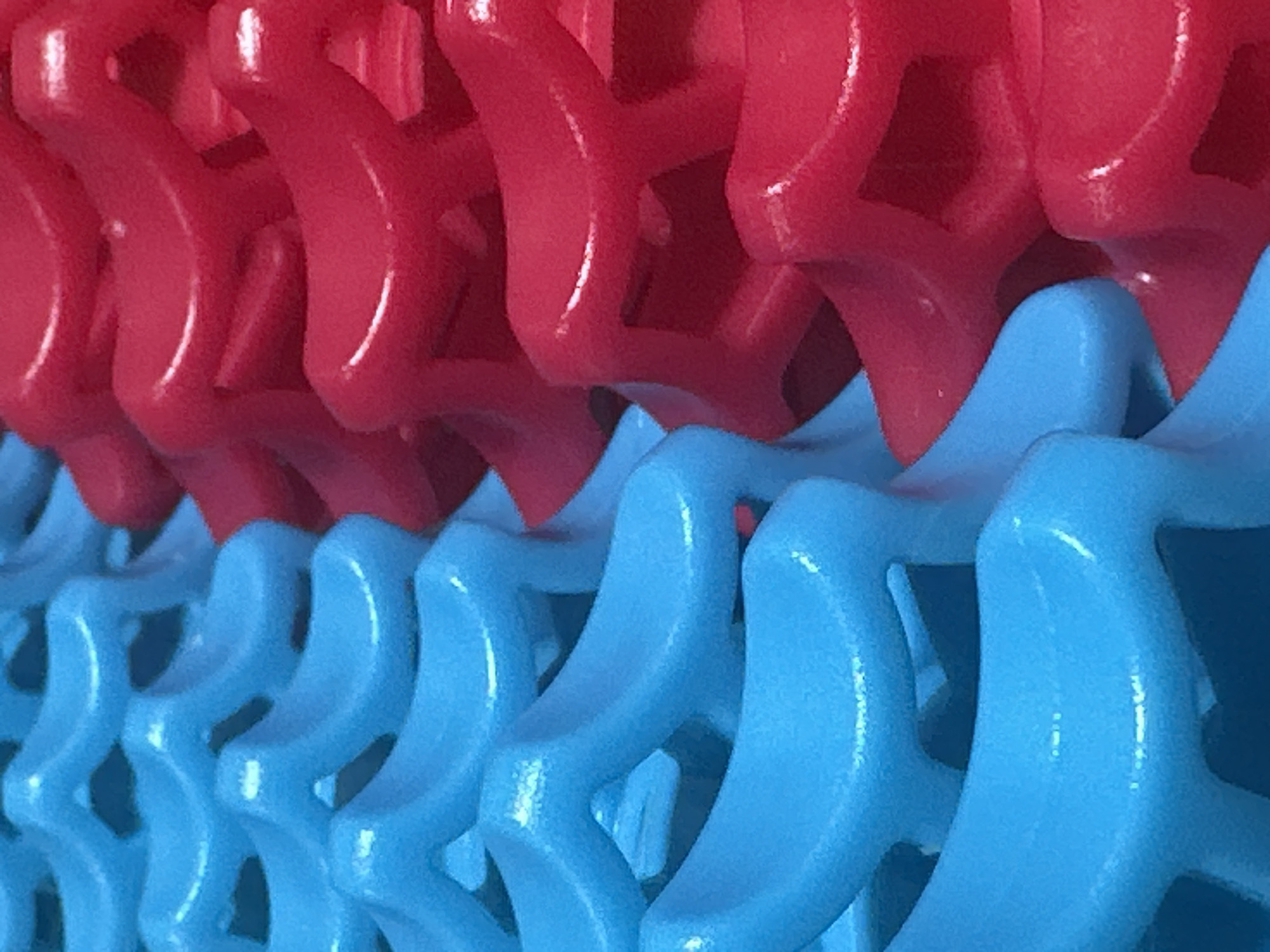 The image shows a close-up, abstract view of interlocking blue and red plastic elements, arranged in parallel rows that appear to be part of a larger structure or toy. The texture of the plastic is slightly rough, with subtle highlights indicating a matte finish. The perspective is diagonal, with the blue elements occupying the left side of the frame and the red elements the right, suggesting they are intermeshing or stacked. There are no people or discernible environmental features present in the image; it is solely focused on the abstract pattern of the colored plastic components. There is no information in the image to suggest a location in Letterkenny, Ireland.
