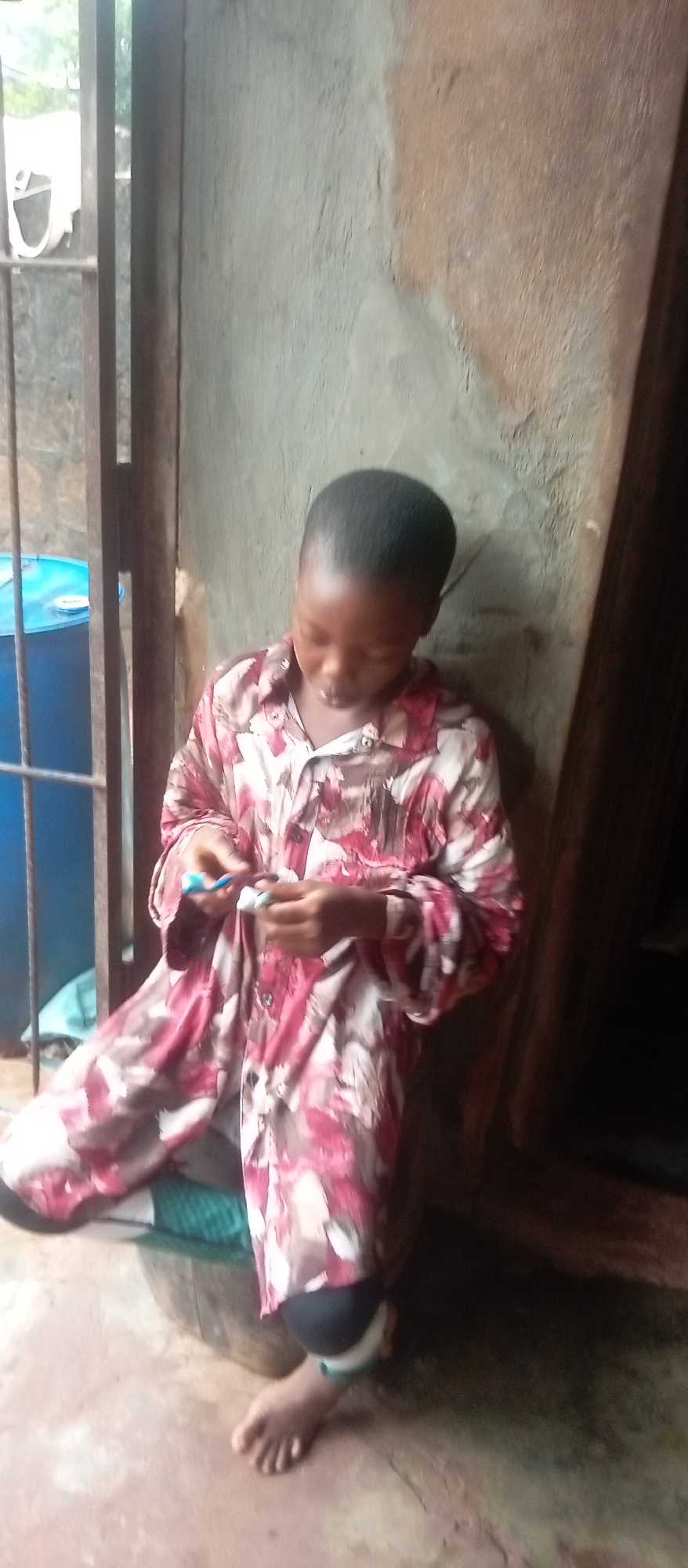 In Ogidi, Nigeria, a young person with short dark hair, wearing a patterned shirt with shades of red, white, and brown, is seated on a wooden barrel. They are looking down at their hands, which are engaged in an action that appears to involve manipulating small objects. The background consists of a rough, textured wall and vertical metal bars. To the left, a blue cylindrical object is visible behind the bars. The ground appears to be made of reddish-brown earth.
