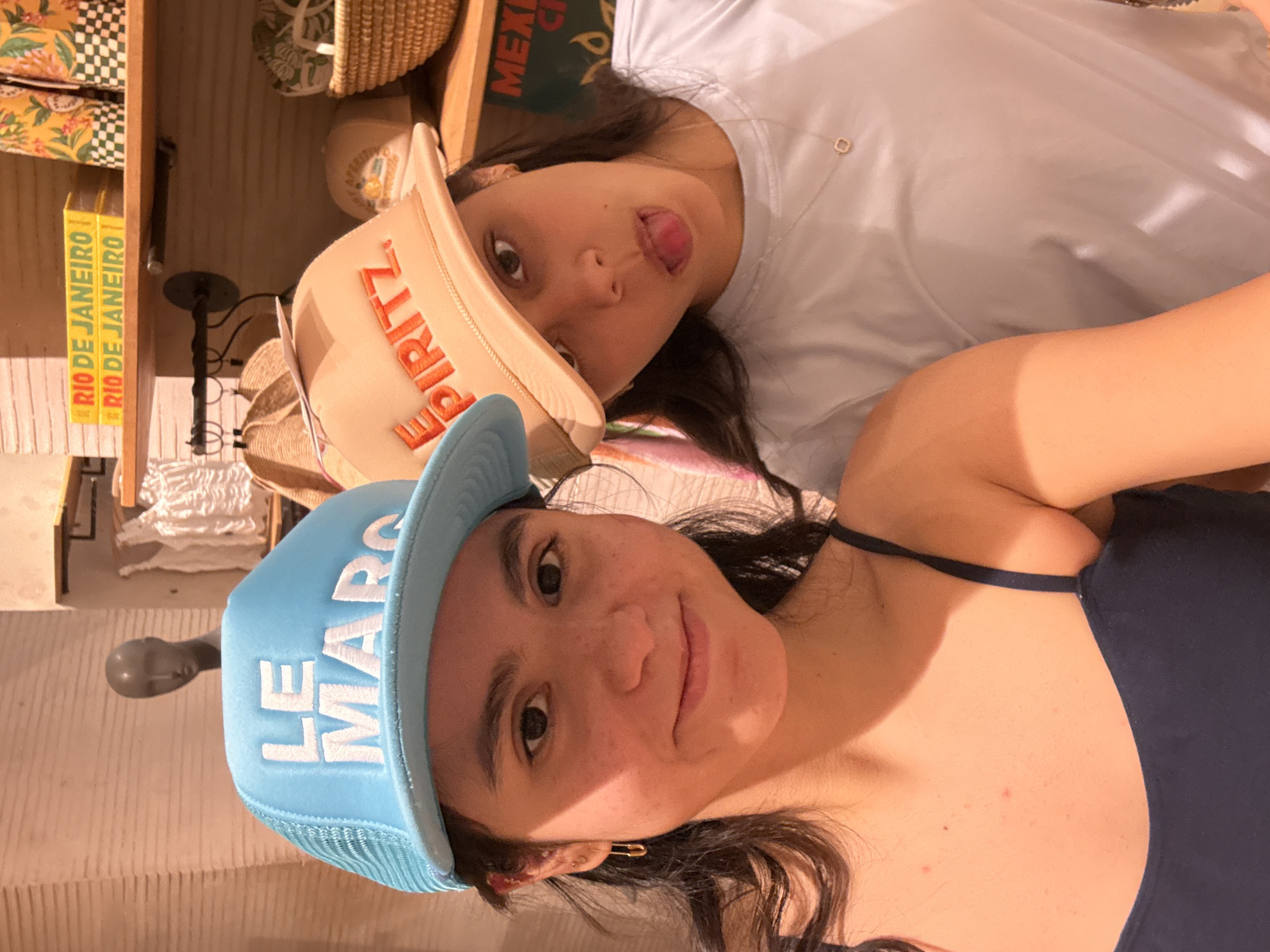 Two young women pose for a close-up photograph in what appears to be a retail setting. The woman in the foreground wears a light blue trucker hat with the words "LE MARCO" in white capital letters. She has dark hair, light skin, and is wearing a dark-colored tank top with thin straps. Her hair is styled with loose waves. She has a small gold safety pin in her ear.
Behind her, another woman wears a light brown hat with the word "SPRITZ" stitched on the front. She has long dark hair and is wearing a white t-shirt. She has her tongue sticking out.
Behind the women, there are shelves containing various items, including hats, books, and bags. "RIO DE JANEIRO" is printed on the spine of several books. The environment suggests an upscale retail store, but the specific location is not visually identifiable.