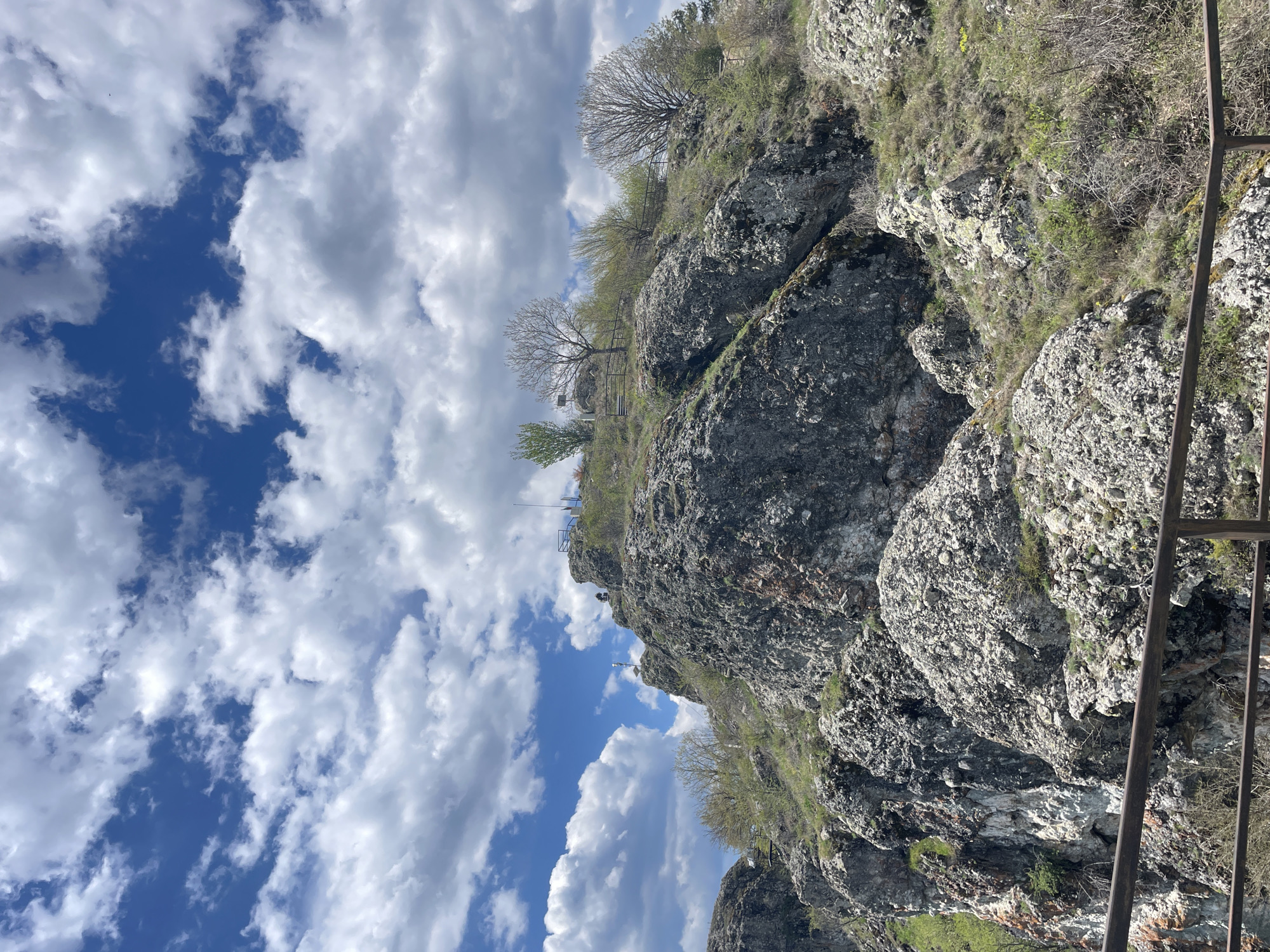 The image shows a rocky cliffside with sparse vegetation, under a sky filled with cumulus clouds. The sky exhibits patches of blue. The cliff face is composed of rough, grey rocks. Greenery is visible on the cliff's edges.
A metal railing appears on the right side of the frame, suggesting a constructed viewpoint or path. On top of the cliff, there are small, bare trees and what appears to be a metal safety barrier.
The location is Gradat, Bulgaria.
