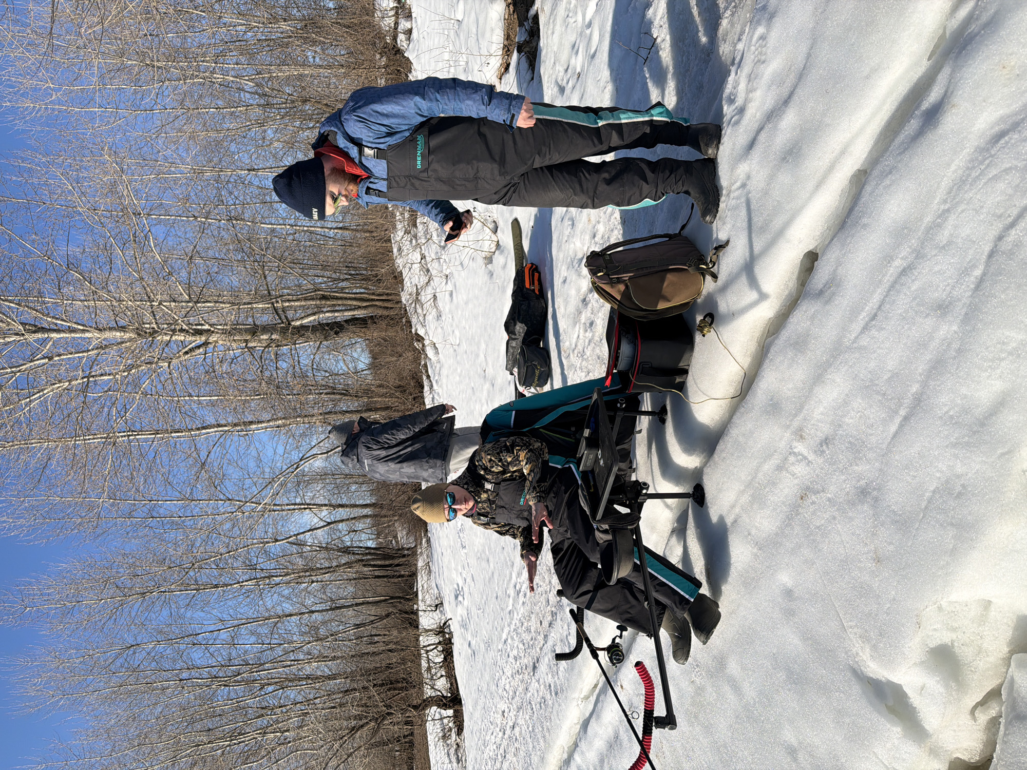 Two people are ice fishing. One person is standing, wearing a blue jacket, black bib overalls with turquoise accents, a black hat, and sunglasses. This person is holding a dark object in their hand and looking down. The other person is seated in a fishing chair. The chair has attachments for fishing rods and a small table. The seated person is wearing a camouflage jacket, black bib overalls, a beanie, and sunglasses. Fishing gear, including bags and fishing rods, are scattered on the snow. The background shows a snowy landscape with bare trees and a bright blue sky. The location is in a snowy, possibly forested area within the city of Chaikovsky, Russia.
