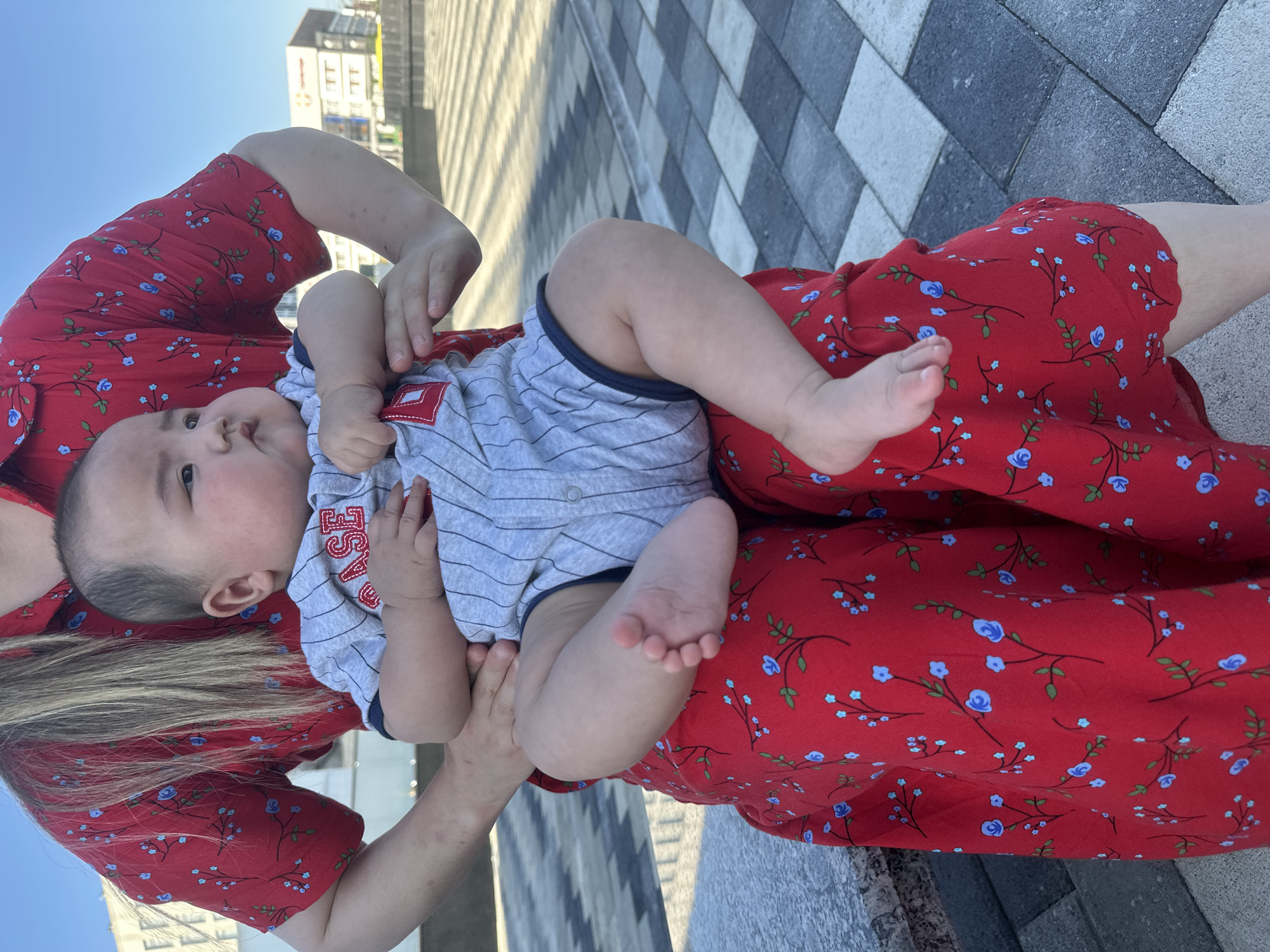 A close-up shot features a baby being held by a person. The baby, wearing a grey striped onesie with red lettering that appears to say "BASE", is looking towards the camera. The person holding the baby is wearing a red dress with a floral pattern of small blue flowers and green leaves. The background shows a pathway made of grey and white rectangular tiles, with sunlight casting diagonal shadows across them. In the upper portion of the image, a building with multiple windows is visible against a clear blue sky. The scene appears to be outdoors, possibly in a city setting given the building and tiled walkway. The lighting suggests it is daytime, likely sunny. The overall impression is a tender moment between a caregiver and a baby.