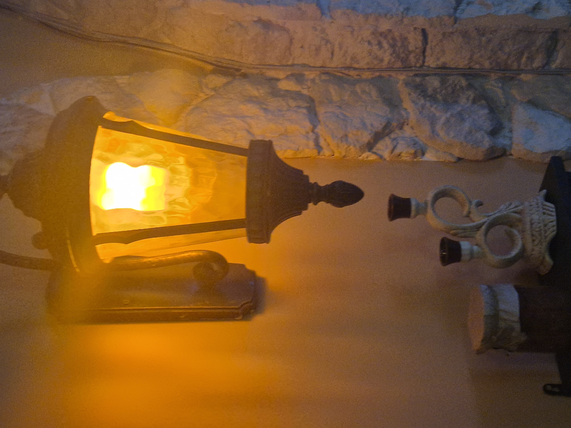 An ornate, vintage-style wall lantern with a glowing yellow light hangs on an orange-toned wall. To the right, a textured stone wall features an arched alcove, illuminated by the lantern's warm glow. In the foreground, a dark shelf holds two objects: a cylindrical jar with a textured lid and a decorative white candle holder with two prongs for candles. The candle holder has an intricate, swirling design. The scene appears to be indoors, possibly in a home or a restaurant, with a warm and cozy ambiance. The location in Kokshetau, Kazakhstan is not determinable from the image.