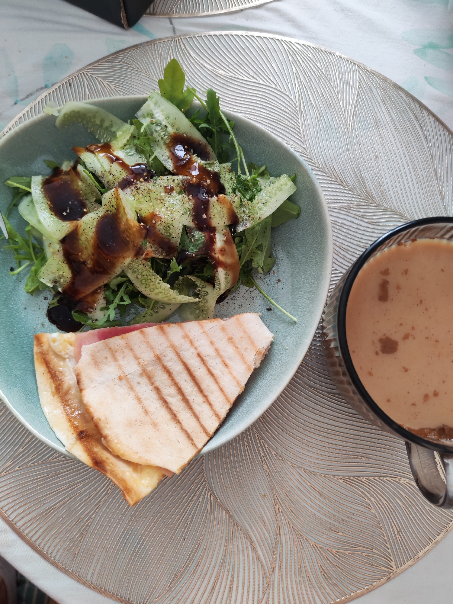 The image displays a close-up, overhead view of a meal on a decorative placemat. In the foreground, a pale blue bowl contains a salad with thinly sliced cucumbers, green leaves, and a dark drizzle of dressing. Beside the salad, a grilled flatbread sandwich, cut in half, is visible, revealing a slice of pink ham and melted cheese. To the right of the bowl, a clear glass mug holds a creamy, light brown beverage with some small dark flecks. The meal is set on a textured placemat with an intricate, feathery pattern. The background is partially obscured, showing a white and light blue patterned surface. There are no people visible in the image. The scene appears to be a casual dining setting, possibly a home or a cafe. The location is not specified in the image, and therefore, it cannot be definitively linked to Izhevsk, Russia.