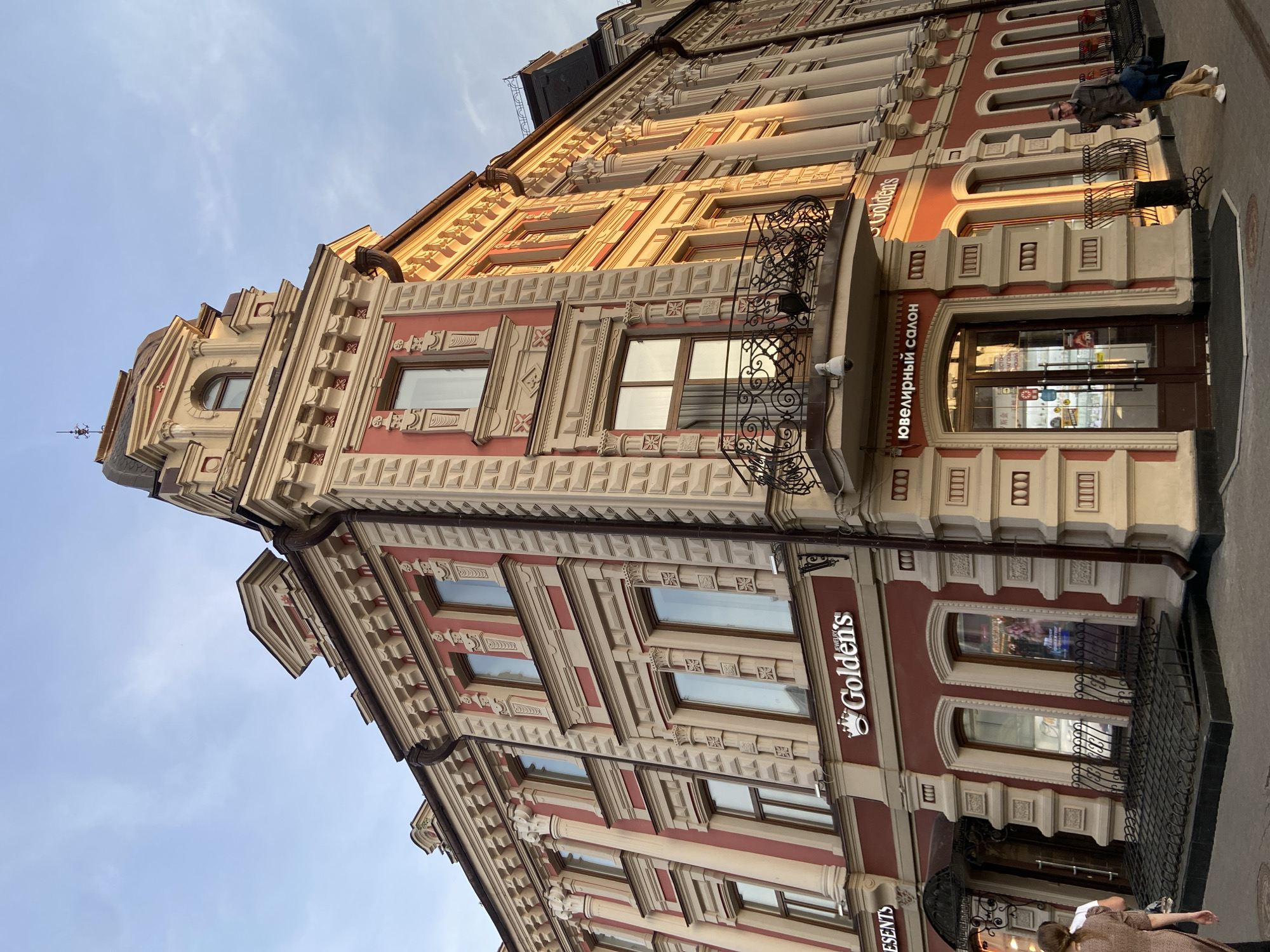 The image shows a building with ornate architectural details. The building is predominantly red and beige, with multiple windows and decorative trim. It appears to house commercial establishments, as indicated by signage in Russian and English: "Ювелирный салон" (Jewelry Salon) and "Golden's" are visible. A wrought iron balcony is present on one of the upper floors. The photo is taken from a low angle, looking upward at the building. It is located in Kazan, Russia.