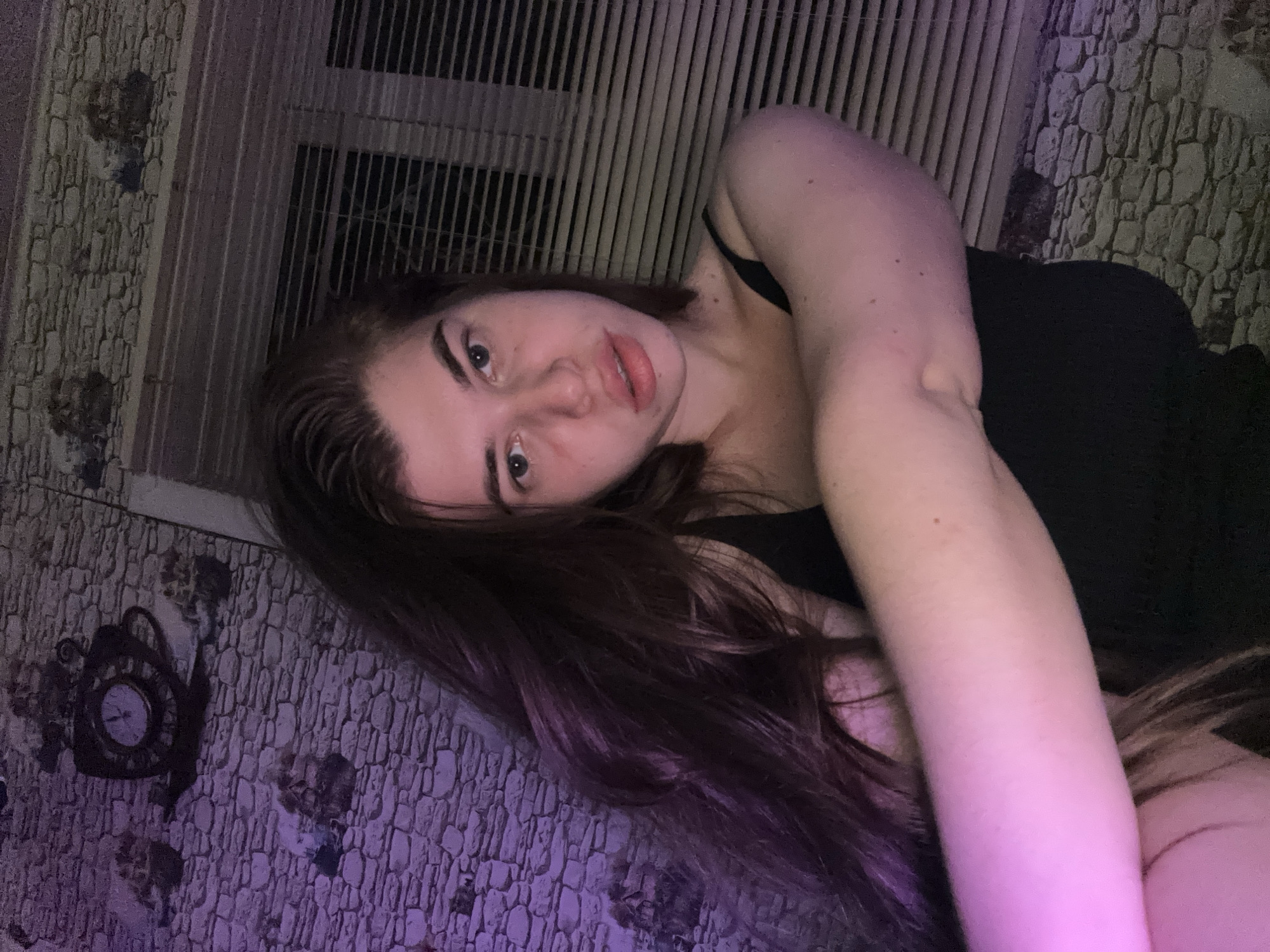 A young woman with long, dark hair is captured in a selfie taken indoors. She is wearing a black tank top and is positioned in the foreground, with her face and upper body prominently featured. Her hair is falling over her shoulder, and her arm is resting across her chest. The background consists of a wall with a stone pattern wallpaper and a window covered by horizontal blinds. A purple light casts a glow on the left side of the image, illuminating her hair and arm. The overall impression is that of a casual, personal photograph, likely taken in a residential setting. There is no information available to suggest this location is in the city of Мещёры, Russia.