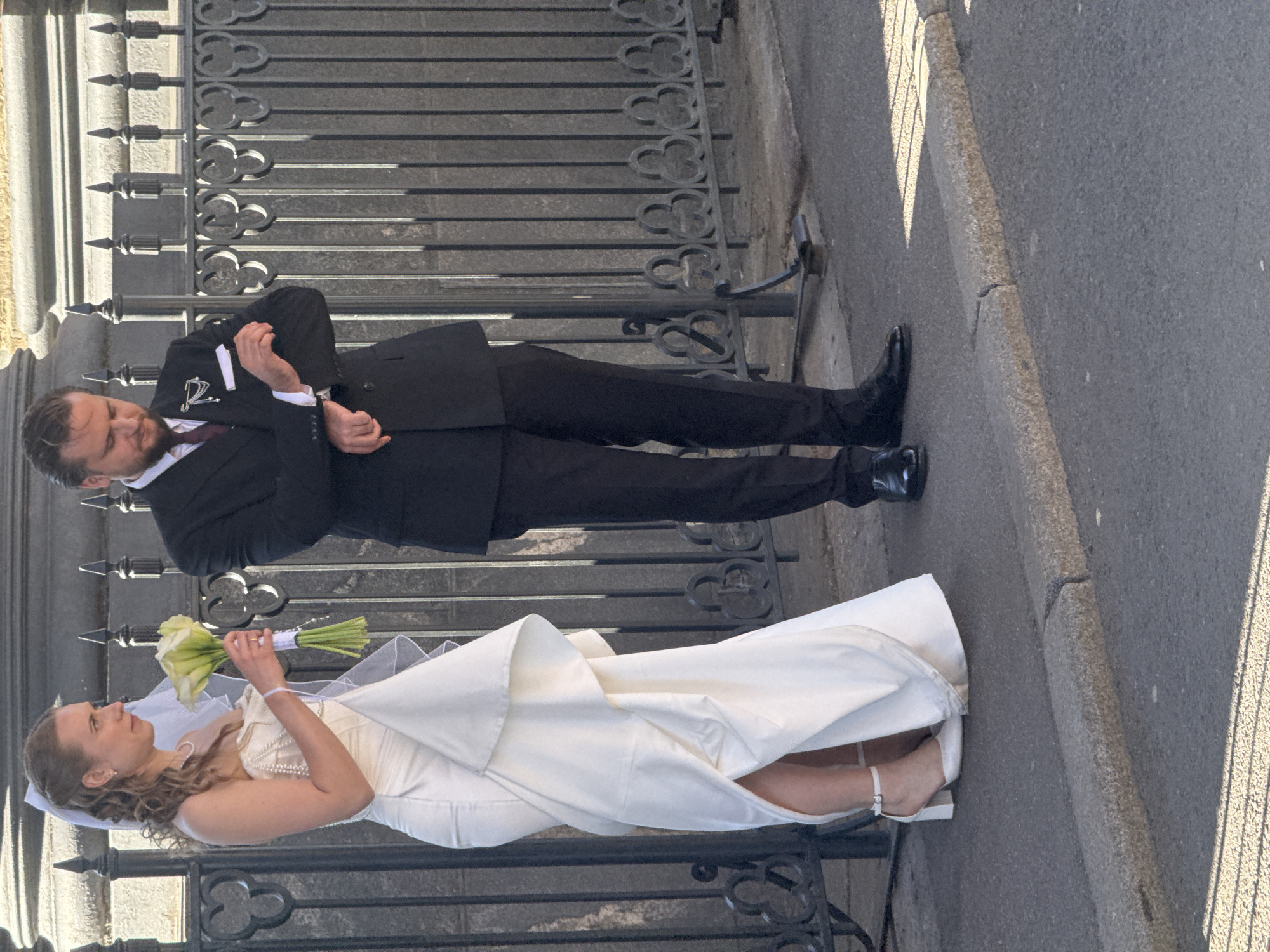 A bride and groom are standing outdoors in front of a decorative metal fence. The bride, on the left, wears a strapless white wedding dress with a draped skirt and veil. She holds a bouquet of white calla lilies and wears white heeled sandals. The groom, on the right, is in a black suit with a dark tie and a pocket square. He is looking down at his wrist, possibly checking a watch. The fence appears to be part of a historical building's exterior, possibly located in Saint Petersburg, Russia, given the architectural style of the fence and the bright sunlight suggesting a temperate climate. The ground is paved with asphalt and a granite curb is visible in the foreground.