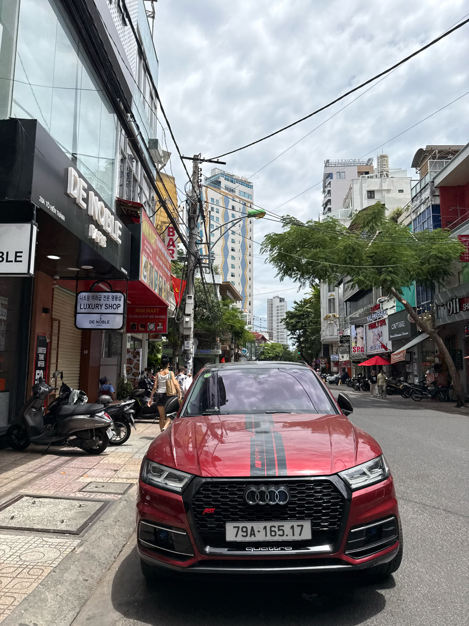 This is a street-level, eye-level shot of a red Audi Q5 parked on the right side of a city street in Nha Trang, Vietnam. The car is prominently featured, taking up the lower half of the frame. Its license plate reads "79A-165.17." A stripe decal runs down the hood. To the left of the car, a sidewalk is lined with shops. A dark-colored scooter is parked on the sidewalk. Further left, a building with a large glass facade and the signage "DE NOBLE PARIS" is visible. A woman in a white tank top and dark shorts walks away from the camera on the sidewalk. The street itself has other parked scooters and a few pedestrians in the background. Buildings of varying heights line the street, with overhead electrical wires crisscrossing the scene. The sky is overcast.