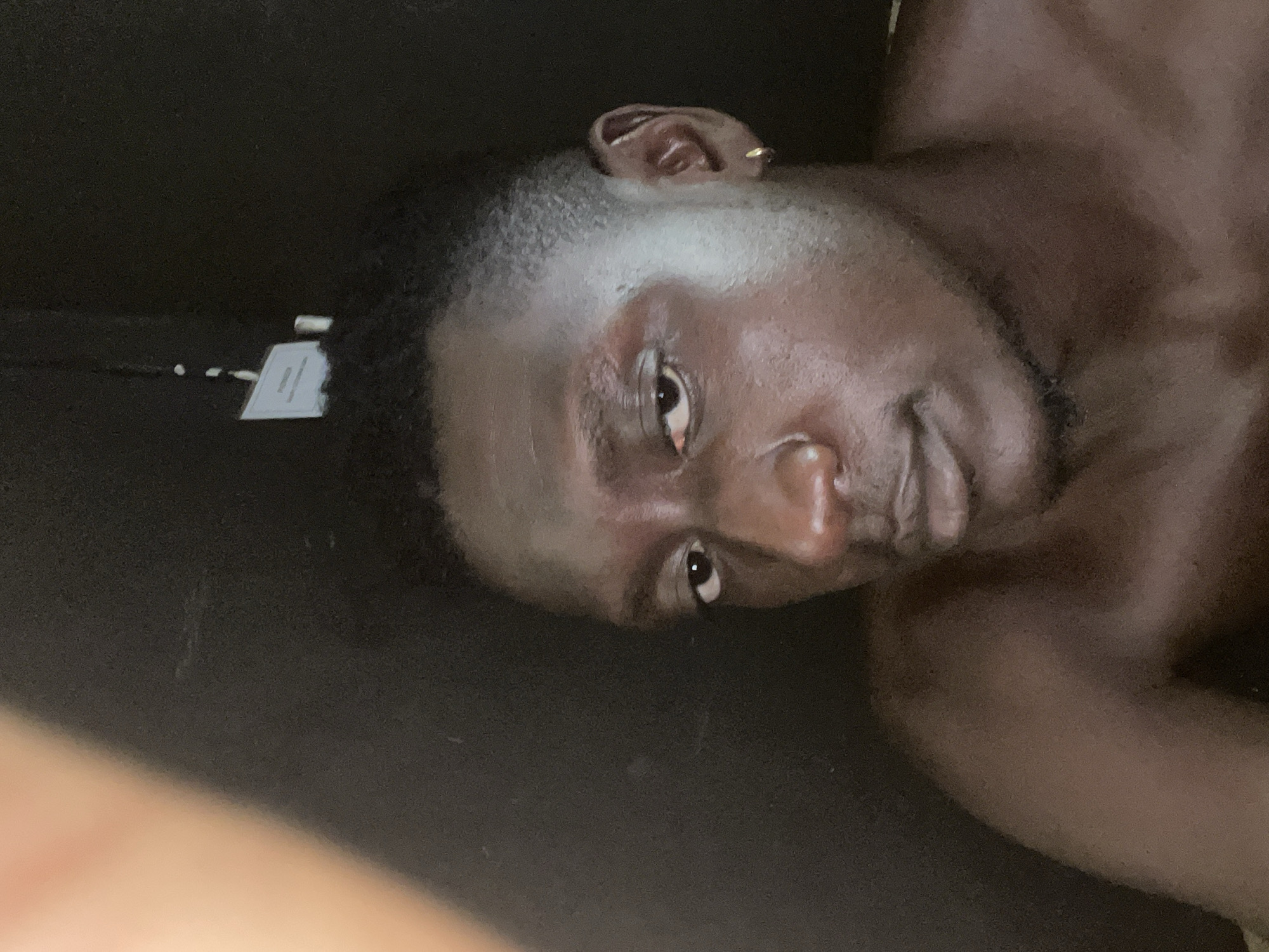 A close-up, angled portrait of a dark-skinned man with short, graying hair. He is shirtless. He has a gold earring in his left ear. A small, white tag with a black cord is visible to the left of his head. The background is a dark, solid color. There is no identifiable location in the city of Jos, Nigeria.