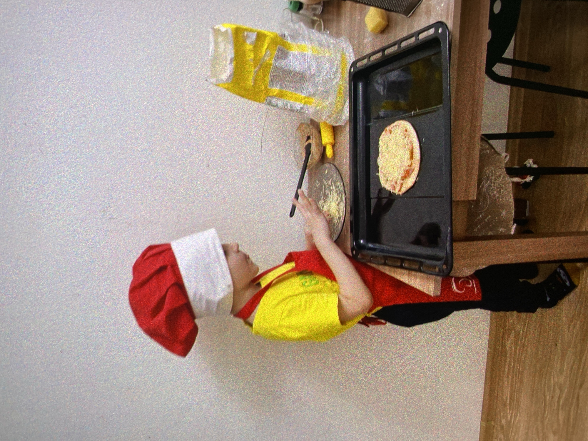 This media file captures a young child dressed as a chef, preparing food in an indoor kitchen setting. The image is oriented vertically, showing the child from the chest up, standing or seated at a wooden counter.
The main subject is a child wearing a red and white chef's hat, a bright yellow t-shirt, and a red apron. Their hands are positioned over a clear glass plate containing grated cheese or a similar topping, with a black spatula resting on the plate. Next to the plate, on a black baking tray, lies a small, round, unbaked pizza or flatbread, partially covered with toppings.
The immediate surroundings on the wooden counter include a large, crinkled yellow bag, likely containing food ingredients such as flour, and a grater. A dark chair is partially visible in the background to the right. The wall behind the child is a light, speckled off-white color. The scene suggests an activity of assembling a pizza or similar baked dish.
The image appears to be taken in a well-lit environment, presumably during the daytime, though no windows are visible. The overall texture of the image is somewhat grainy. There is no clearly legible text on any visible objects, although faint designs or text might be present on the yellow bag, the child's yellow t-shirt (possibly green text), and the red apron (possibly white design). The location context of Saint Petersburg, Russia, is not visually discernable from the image content.