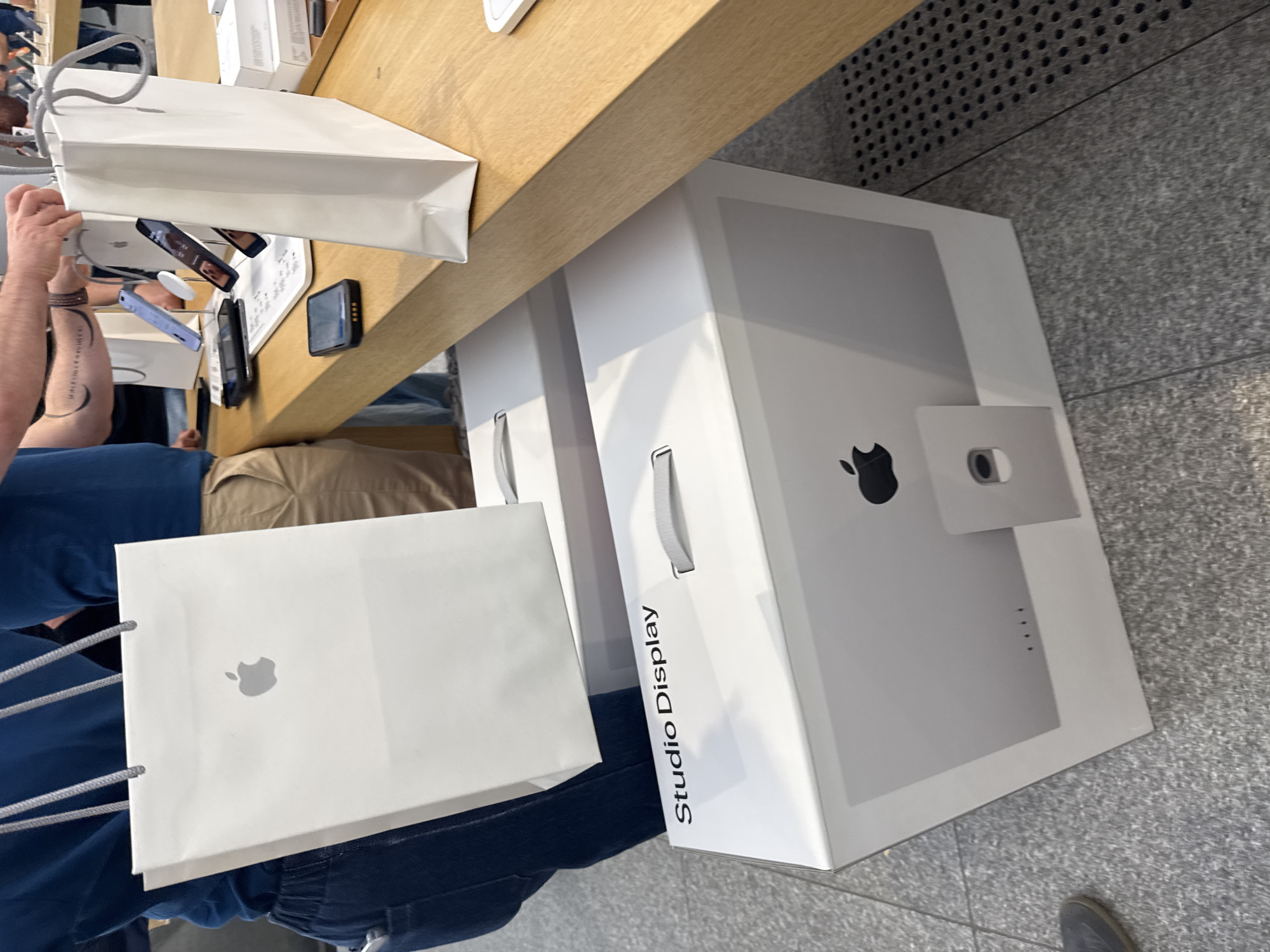 The image captures a scene likely within an Apple Store, indicated by the presence of Apple shopping bags and boxes of Studio Displays. An individual, visible from the mid-torso down, is holding an Apple shopping bag with the Apple logo prominently displayed. A second person is holding an additional white Apple shopping bag with gray handles. Next to this bag is the packaging for an Apple Studio Display. A portion of the display itself is visible on the box. The setting appears to be a retail environment, with a wooden display table holding various electronic devices, including smartphones and keyboards. The floor is a typical light-colored, possibly concrete or tile surface, found in many retail spaces. While Milan is mentioned in the instructions, there is no evidence in the image to identify the city. The photograph focuses on the items related to Apple products and the act of shopping in a retail setting.