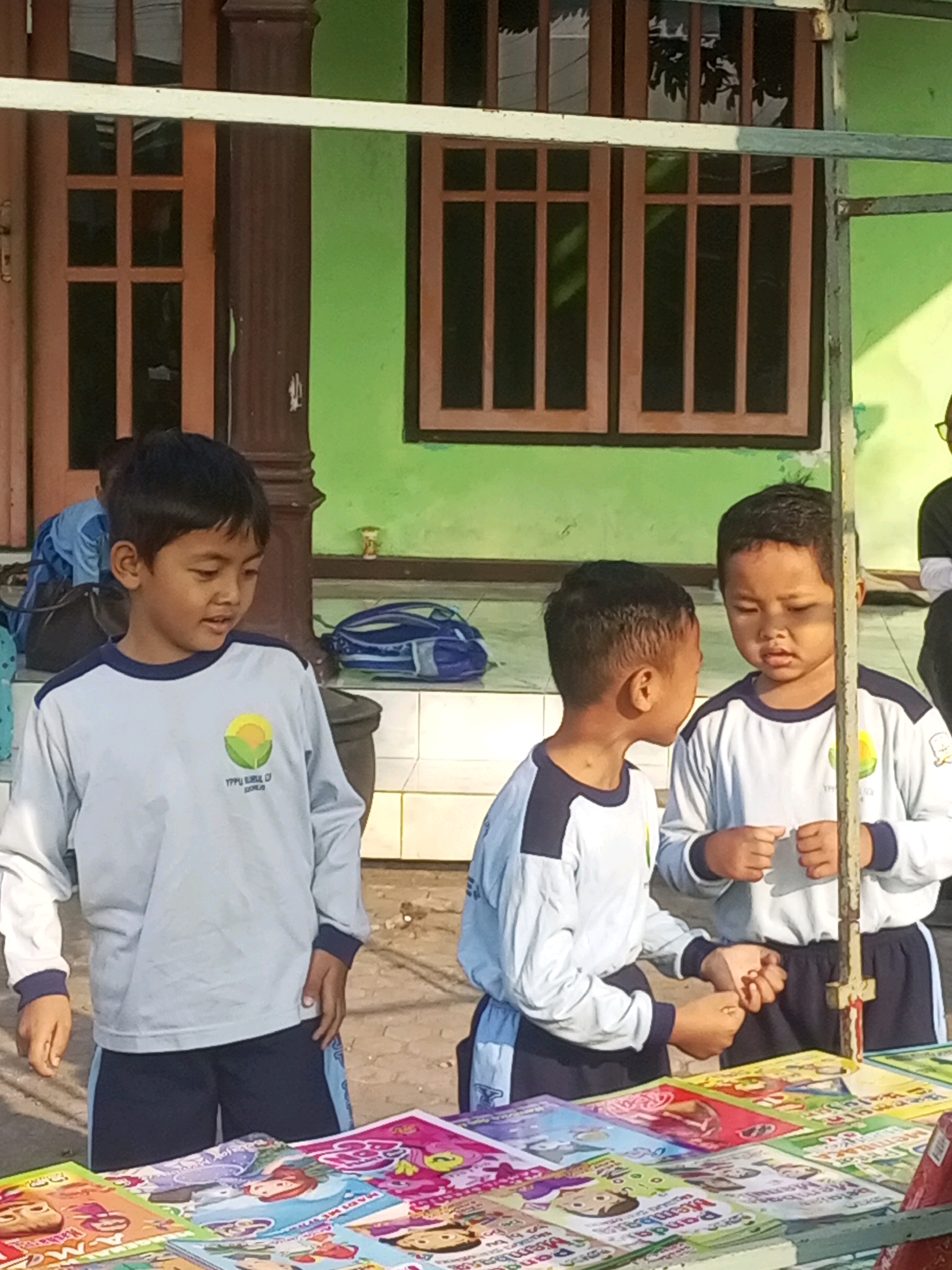 In Gurah, Indonesia, three young boys stand near a table laden with colorful children's books. The boy on the left, wearing a light blue long-sleeved shirt with a dark blue collar and cuffs, and dark blue shorts, looks to his right. The middle boy, also in a similar light blue long-sleeved shirt and dark blue shorts with a light blue stripe, faces the boy on the right, speaking to him. The boy on the right, in the same uniform, looks down at his hands, which are clasped together. Behind the boys, a green wall with wooden-framed windows is visible, suggesting an outdoor setting, possibly near a school or market stall. The overall scene depicts a casual interaction among children browsing or presenting books.