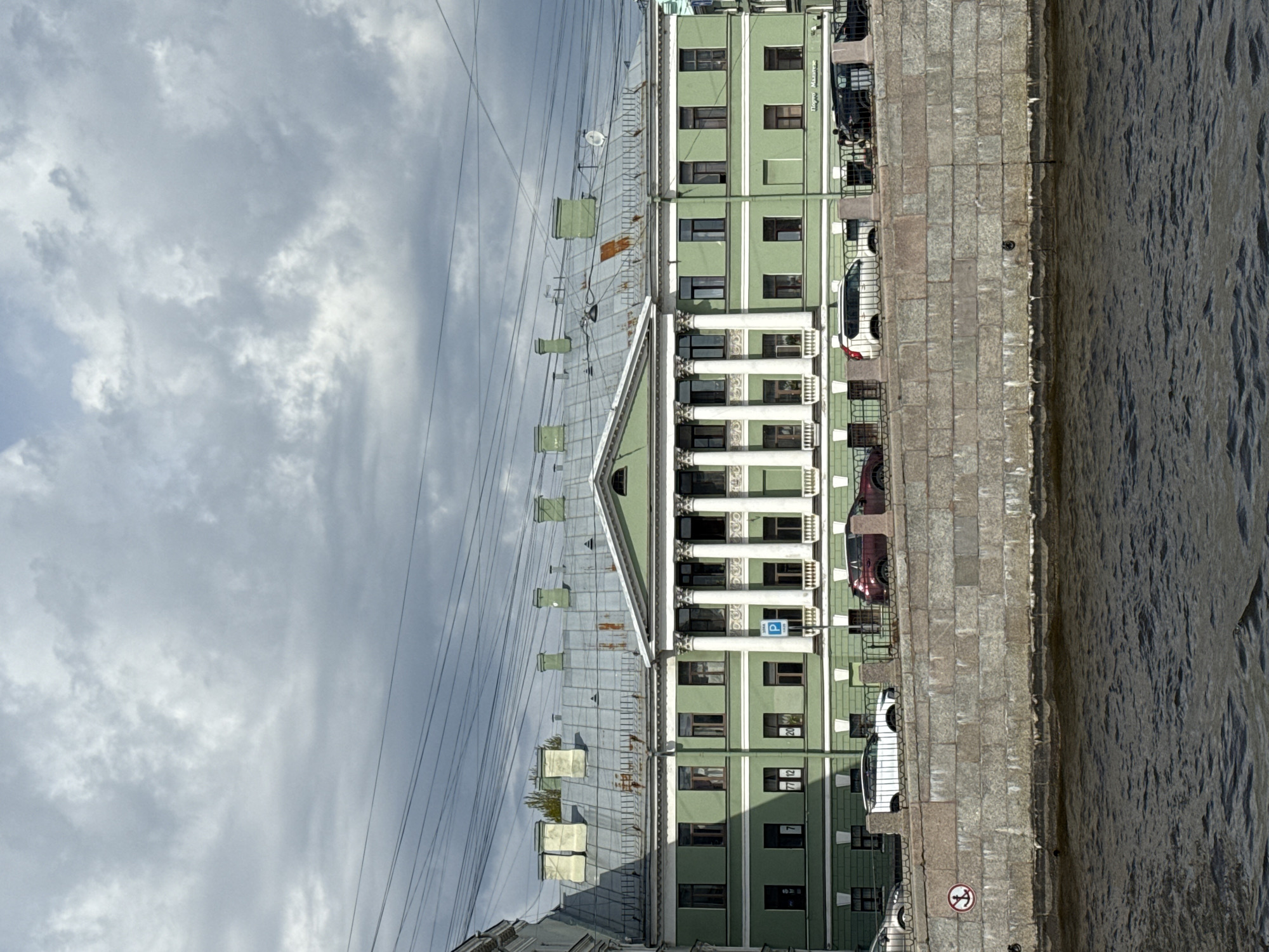 The image captures a street scene in Saint Petersburg, Russia, featuring a green-colored building with white classical columns, situated along a stone embankment next to a body of water. Several parked cars are visible along the embankment. Overhead, a network of cables and wires stretches across the sky, which is filled with overcast, cloudy conditions. The building appears to be a historical structure, possibly a governmental or cultural institution, given its architectural style. There are no people visible in the shot. The perspective suggests the photo was taken from a slightly elevated position, possibly from across the water or from a higher floor in an adjacent building.