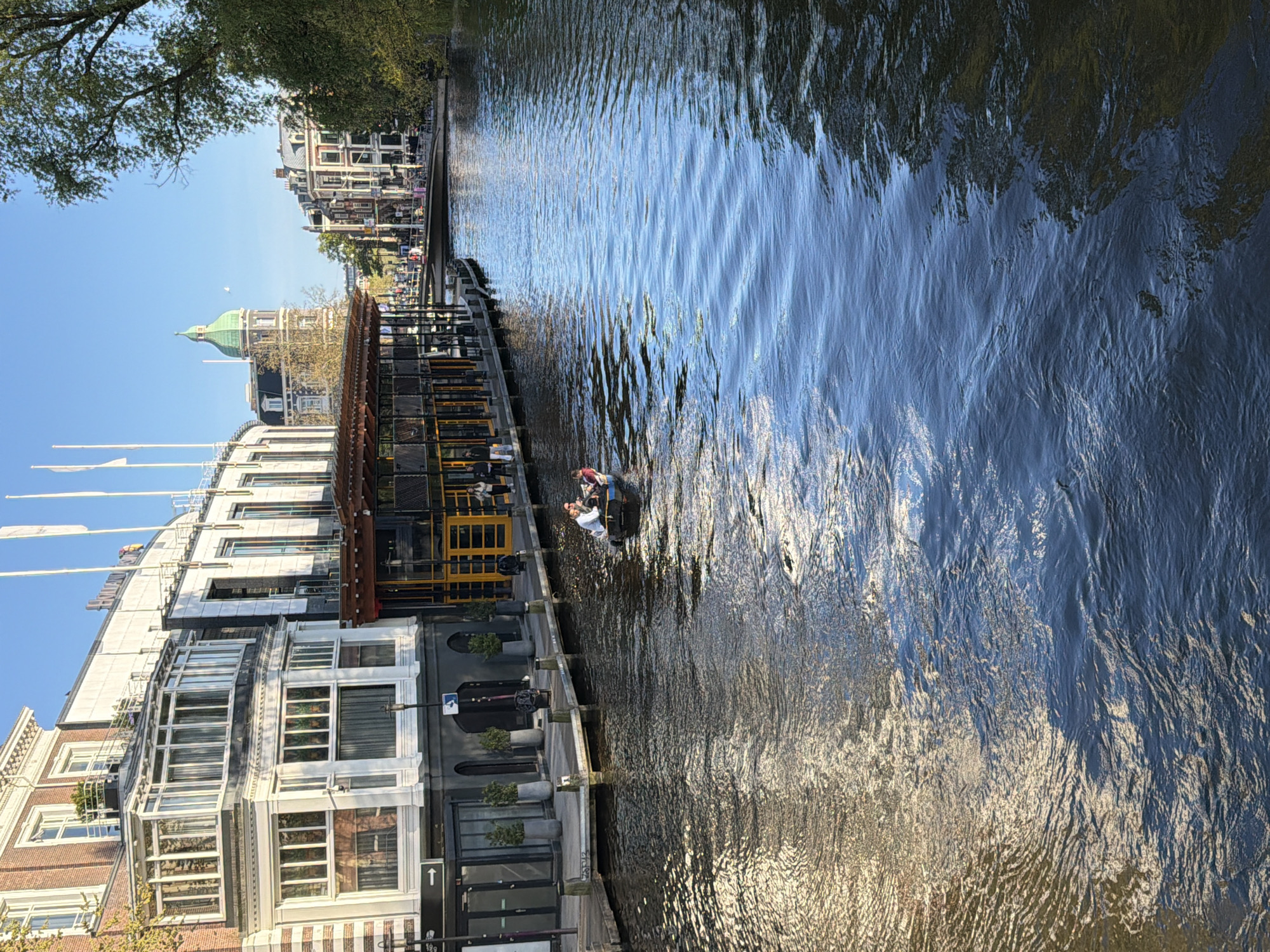 This media file presents a bright, sunny daytime scene along a canal in Amsterdam, Netherlands, viewed from a slightly elevated perspective. The original image is rotated 90 degrees clockwise, but when correctly oriented, it depicts a canal flowing from the foreground to the background, flanked by buildings on its left bank.
The main subjects include people in a small boat on the canal and people seated in a canal-side establishment. The boat, a small open vessel, carries at least two individuals; one appears to be steering from the stern, while another is seated facing them. The canal water is calm yet shows ripples, reflecting the clear blue sky and sunlight, creating patterns of light and shadow.
On the left bank, a variety of architectural styles line the canal. In the background, a distinctive building with a prominent green domed roof and spire rises above the other structures. Closer to the foreground, a modern building with multiple large windows and a white facade is visible, adorned with white flags on its roof. Adjacent to this, an enclosed structure with a dark glass front and a brown roof extends along the canal, appearing to be a restaurant or bar, with patrons visible seated inside. Potted plants and street furniture are also present along this bank. Green tree foliage frames the upper left corner of the scene against a clear blue sky.
The activity taking place involves recreational boating on the canal and people enjoying the canal-side amenities. The time of day is clearly daytime, likely mid-morning or afternoon, given the bright sunlight and the reflections on the water. The weather is clear and sunny. A notable detail on the canal bank is a white directional arrow pointing upwards on a black pole. No other visible text is clearly legible in the image.