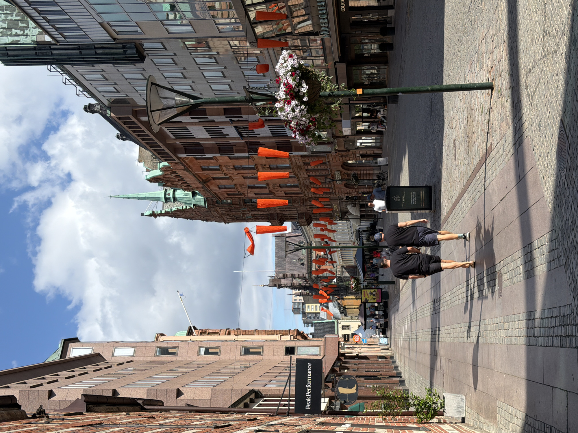 The image depicts a street scene in Malmö, Sweden, specifically the pedestrianized street of Södra Förstadsgatan. In the foreground, two men wearing black t-shirts and shorts walk away from the camera, casting shadows on the cobblestone pavement. A lamppost with a hanging basket of flowers stands to their right, adorned with orange, cylindrical decorations.
The street is lined with multi-story buildings on both sides. The building on the left features a brick facade and a sign for "Peak Performance." The building on the right is characterized by its glass windows reflecting the sky and other buildings, and a sign that reads "SÖDERGATAN." Further down the street, more buildings with intricate architectural details and awnings are visible, along with people and umbrellas suggesting shops and outdoor seating areas. The sky is blue with scattered white clouds.