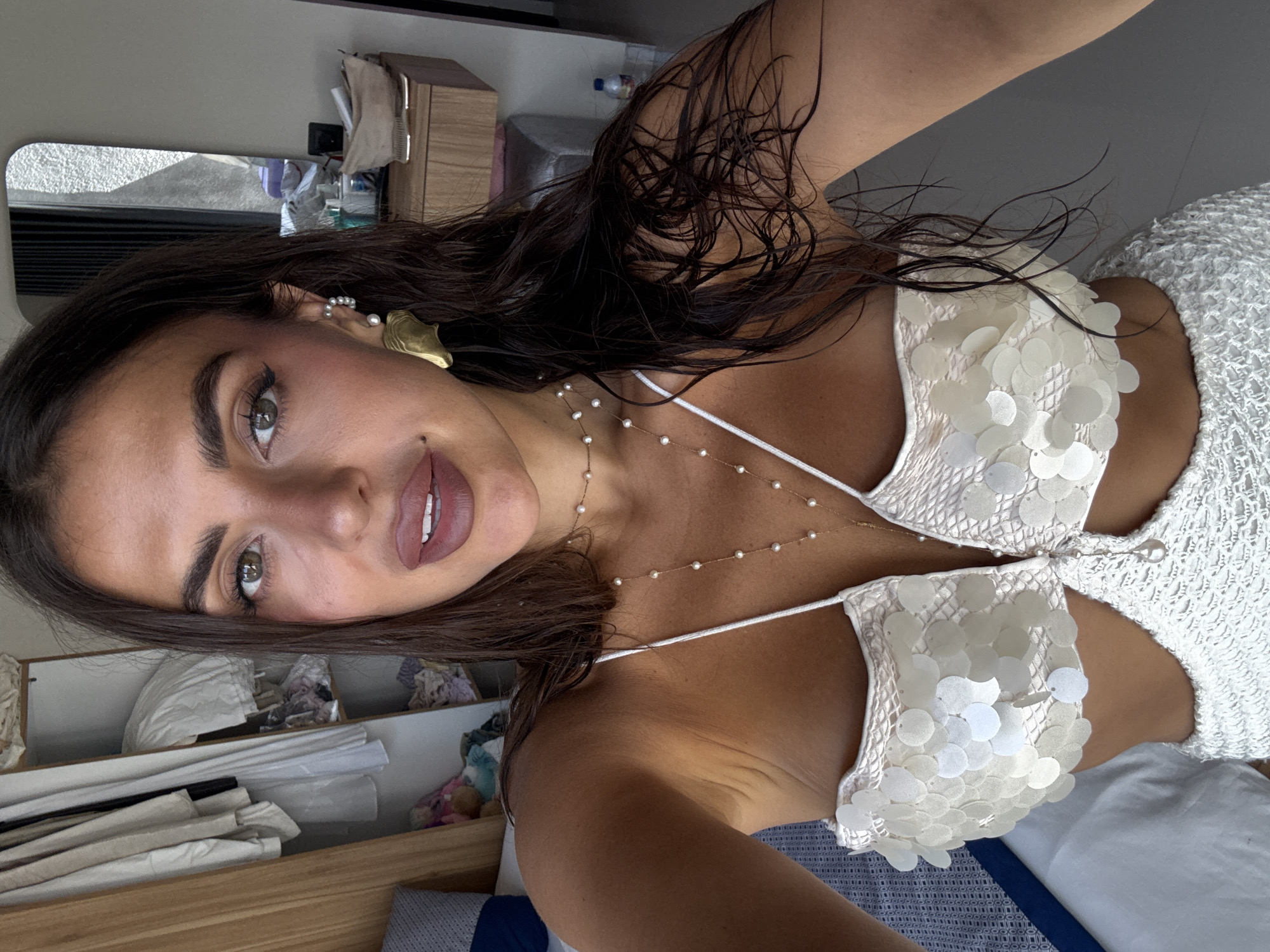 A woman poses indoors, presumably in a private residence in Kuta Selatan, Indonesia. She is facing the camera and partially turned to the side. She has dark hair, brown eyes, and light skin. She wears a white two-piece outfit with circular embellishments and a delicate gold-colored pearl necklace. An earring is visible on the left ear. The background features a bed with blue sheets and folded fabrics on a nearby wooden surface. A mirror and a storage cabinet are partially visible.