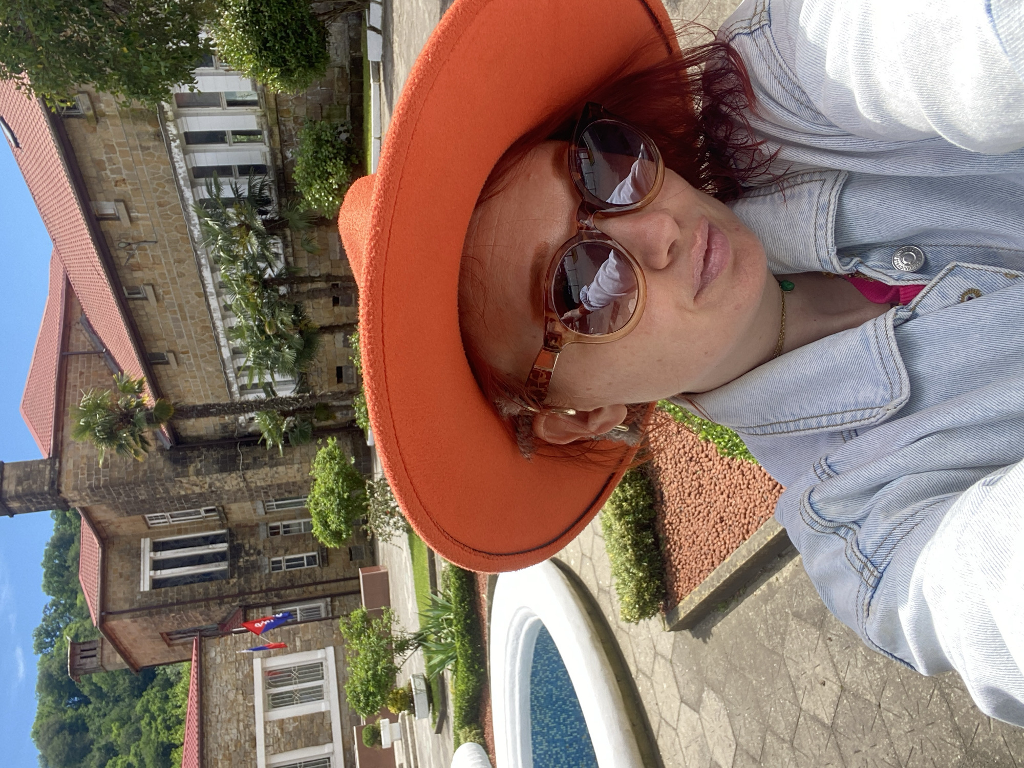 A person is taking a selfie outdoors, with a large building in the background. The person is wearing a bright orange hat, sunglasses, and a light-wash denim jacket. A glimpse of a pink garment is visible under the jacket. The building in the background is multi-storied and constructed of stone. It has a red-tiled roof, and flags are visible on one part of the structure. There are palm trees and other greenery surrounding the building. In the foreground, there is a white fountain with blue tiles. The person and fountain appear to be positioned on a paved walkway. The photo was taken in Sochi, Russia.