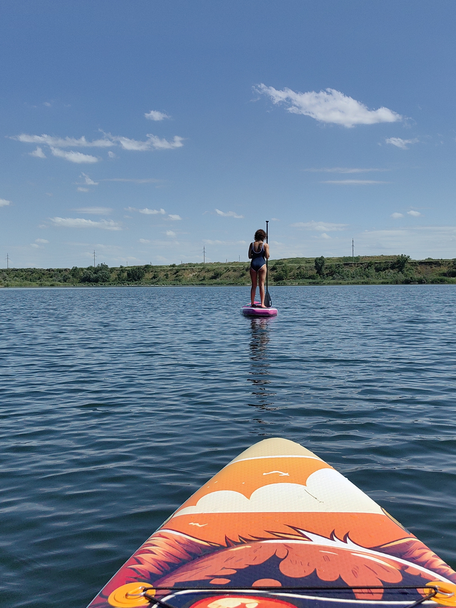 A person stands on a pink paddleboard in the middle of a calm body of water. The person is wearing a dark blue swimsuit and is holding a paddle. The water has small ripples and reflects the blue sky with scattered white clouds. In the background, a tree-lined shore with rolling hills is visible. Electrical poles are scattered along the top of the hills. The foreground shows the front of another paddleboard with an orange and red design featuring a stylized face. The image appears to have been taken on a sunny day in Samoilov, Russia, likely during recreational activity.