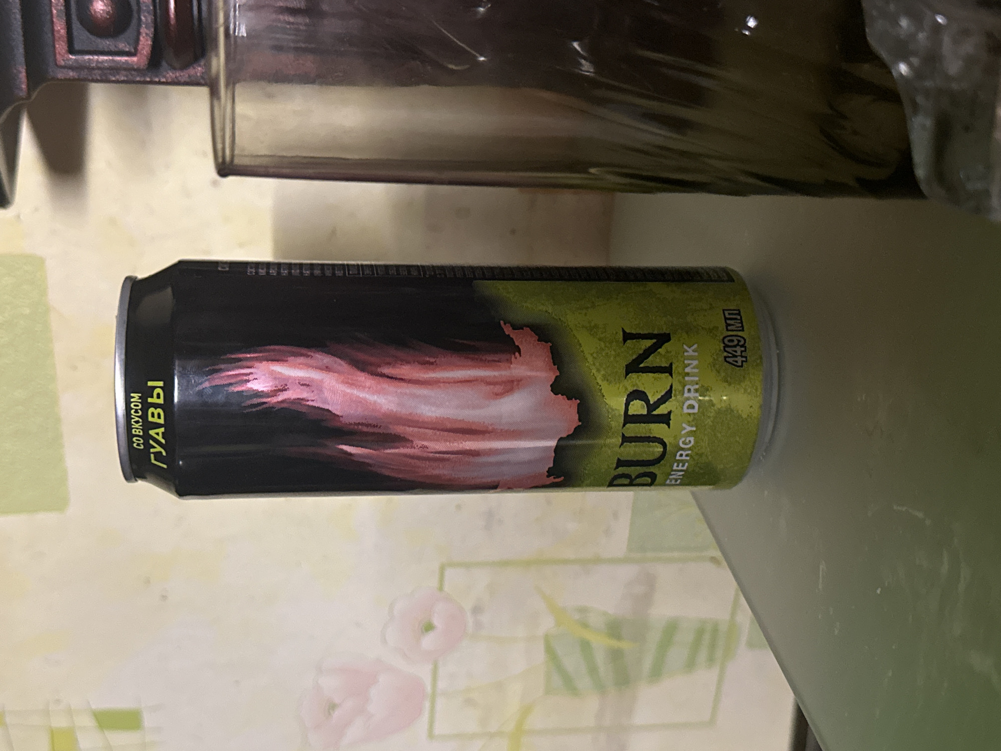A still life photograph shows a can of "Burn" energy drink positioned horizontally on a patterned surface. The can is primarily black with a faded pink design resembling a flame. "Burn" is written in bold, vertically oriented letters on the can, accompanied by the words "Energy Drink." The can indicates a volume of 449 ml. Above the can, out of focus, are parts of other objects which could be metallic. The patterned surface beneath the can has a white or cream base, with floral designs. This is a domestic setting, and due to the Russian writing on the can, the location is Russia, although the city is unknown.