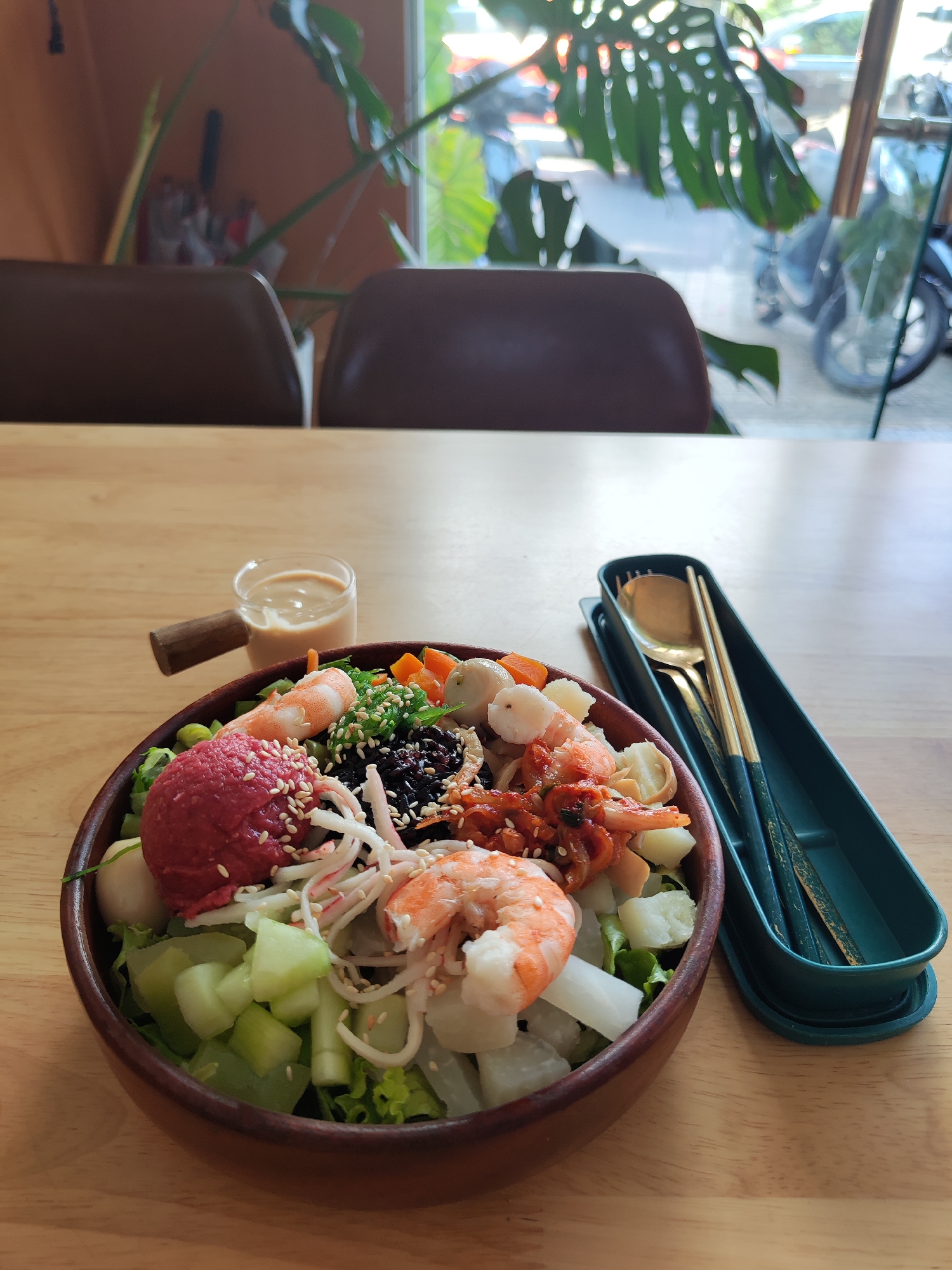 A wooden bowl sits on a wooden table, filled with a colorful mix of ingredients. The bowl contains a scoop of beet-red substance sprinkled with white sesame seeds, shrimp, kimchi, black rice, julienned carrots and lettuce, and sliced cucumbers. A small glass of light brown sauce with a wooden handle is placed to the left of the bowl. A green container with a gold spoon and chopsticks lies to the right of the bowl. Two brown chairs are visible in the background, along with a large monstera plant near a window. Through the window, a street scene is visible. The scene appears to be indoors, possibly in a restaurant or cafe in Da Lat, Vietnam.