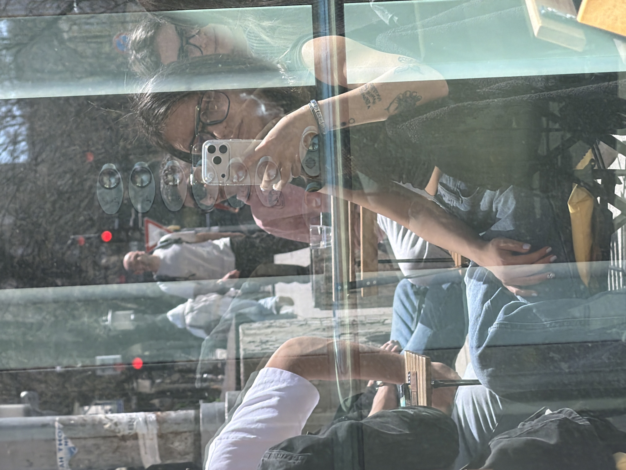The image appears to be a reflection captured on a window or glass surface, creating a composite scene. The primary subject is a person, presumably female, wearing glasses and holding a smartphone up to take a picture. She has dark hair and is wearing a bracelet on her wrist, with tattoos visible on her arm. She is wearing a denim jacket and blue jeans. Behind her and reflected in the glass, a portion of a cityscape is visible, including what seems to be a man walking and street signs with bulbs attached. The building in front has a white facade, and in the background is what looks like buildings and trees. The presence of text-like forms on the glass suggests street signs from the Gabrovo city region of Bulgaria.