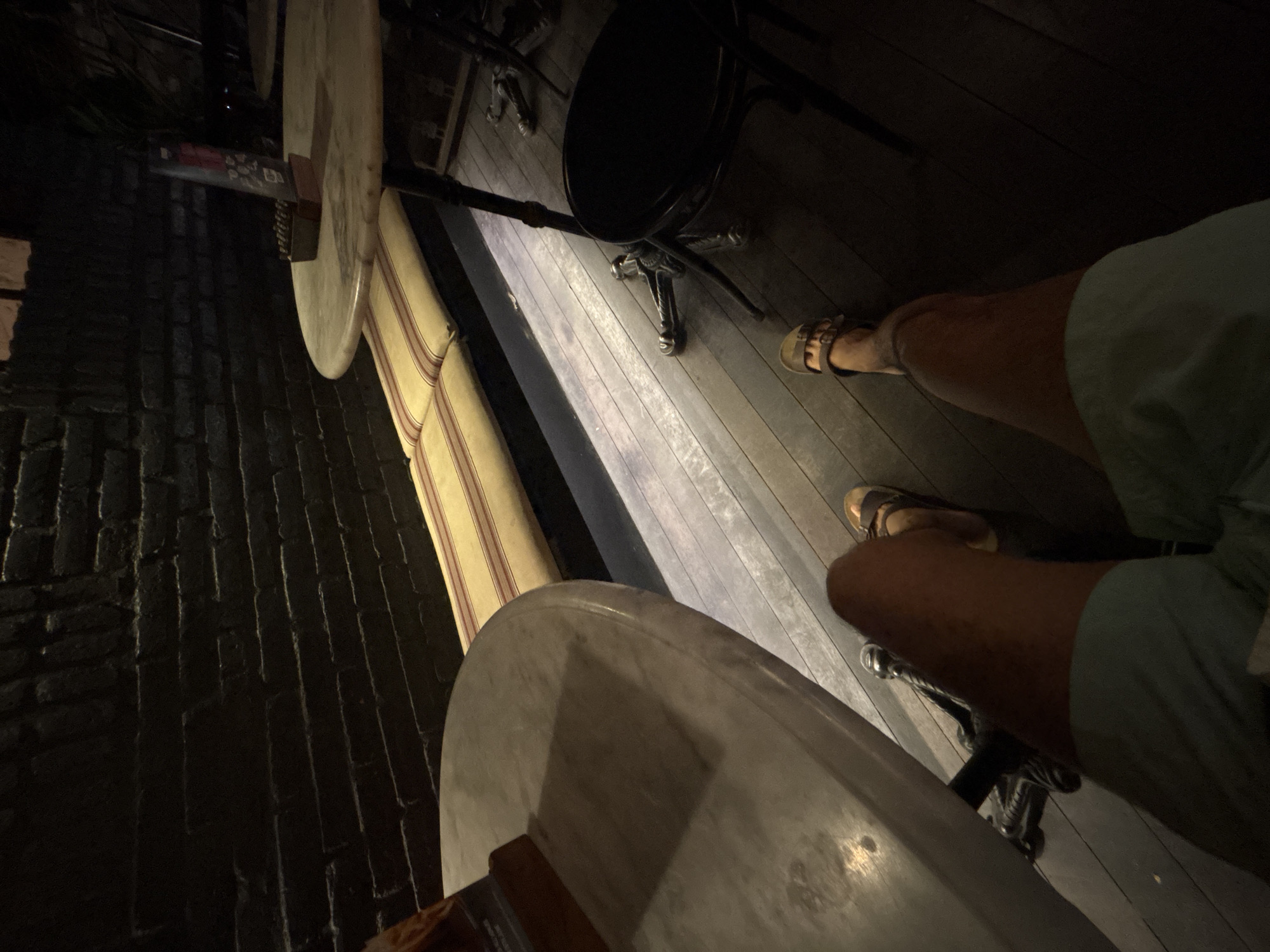 In the city of North Kuta, Indonesia, an overhead shot captures a person's legs and feet, clad in light blue shorts and brown sandals, positioned on a dark wooden floor. The individual is seated at a marble-topped table, its surface partially visible and casting a shadow. To the right, a black stool with a round seat stands near another marble table, topped with a menu and a small holder for what appear to be cigarettes or matches. The background features a dark brick wall and a cushioned bench with yellow and red stripes. Subtle lighting illuminates the floor and the base of the bench, creating a warm ambiance.