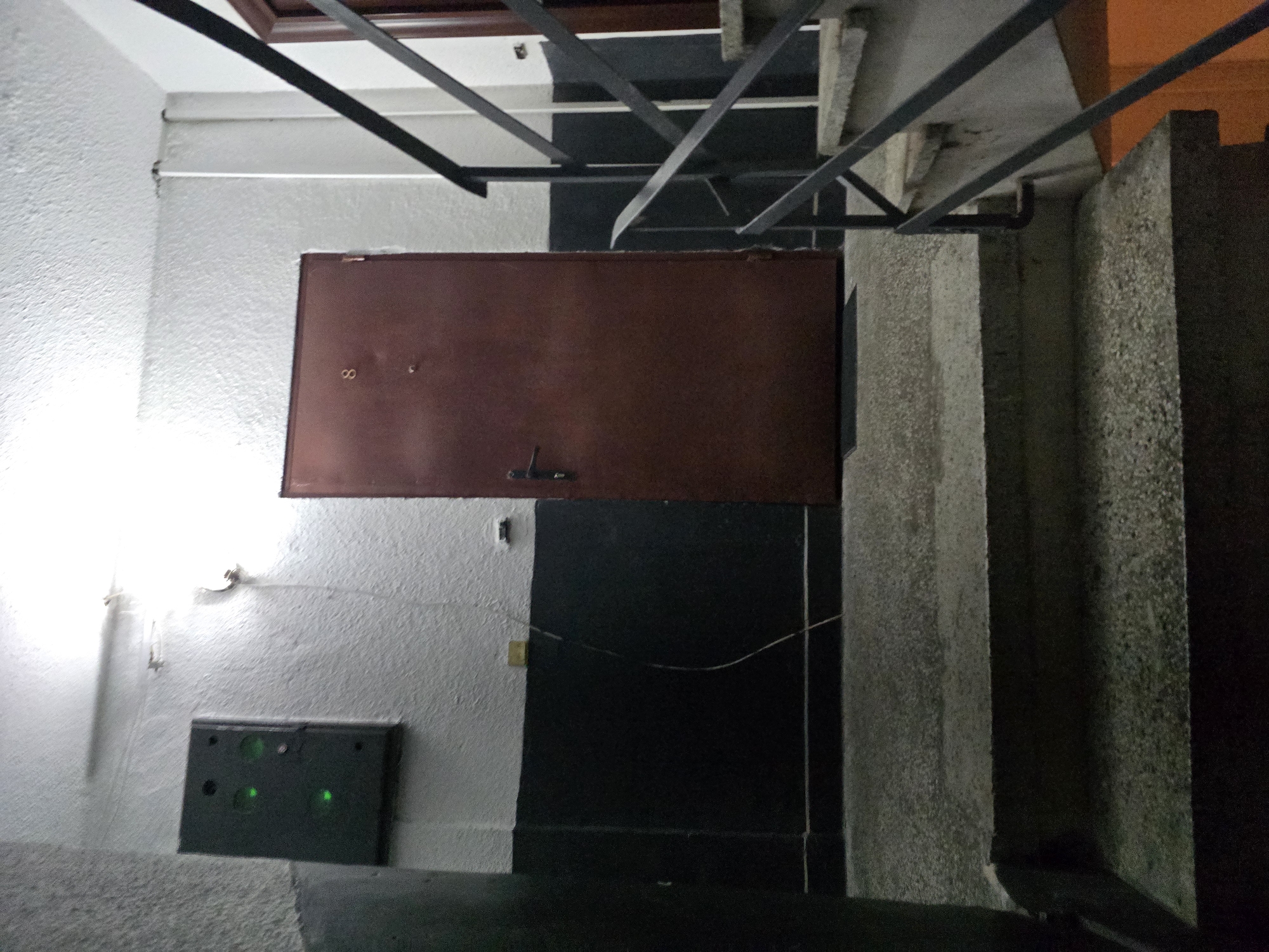 This is a medium shot of a dimly lit interior staircase landing in Yambol, Bulgaria. The foreground shows a set of concrete stairs with speckled texture. A black metal railing runs up the right side of the stairs. The landing area is also concrete. On the left wall, a dark grey rectangular box with three illuminated green lights is mounted. The wall is painted white above a black wainscoting. A dark red metal door, labeled with the numeral "8", is situated on the right side of the landing. To the right of the door, another door frame is visible with a partial view of a dark door. The overall environment appears to be part of a residential building's common area.