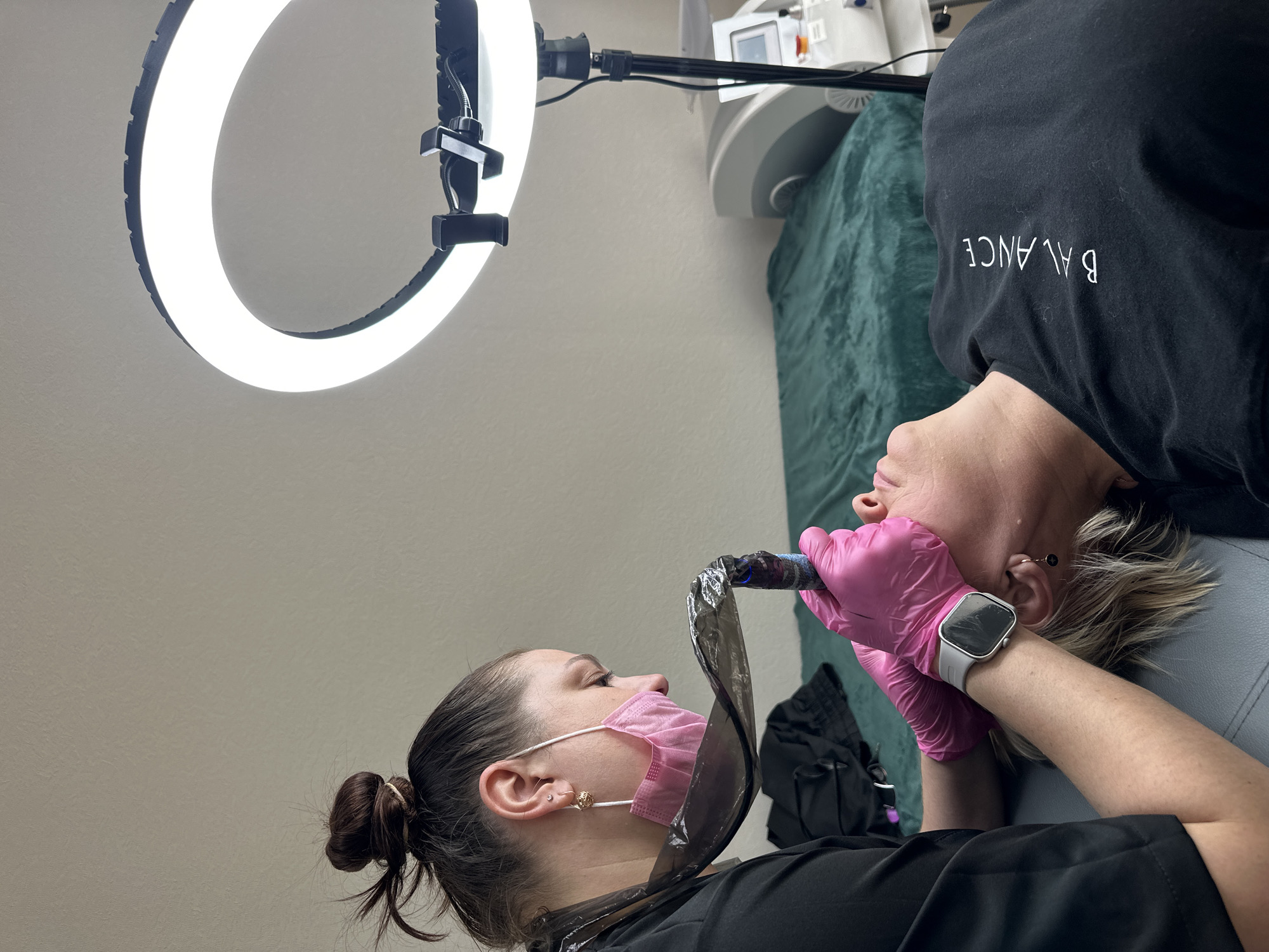 A woman lies on a bed under a large ring light, seemingly undergoing a cosmetic procedure. Another woman wearing a pink face mask, black protective shield, and black attire is positioned nearby, holding a device close to the first woman's face, possibly performing a procedure. Both are indoors in a room with light-colored walls. The location is unspecified but mentioned as Chaikovsky, Russia. Additional equipment is visible in the background, including a machine with a screen. The woman on the bed wears a black shirt with the word "BALANCE" printed in white. The woman performing the procedure wears pink gloves and a smartwatch.