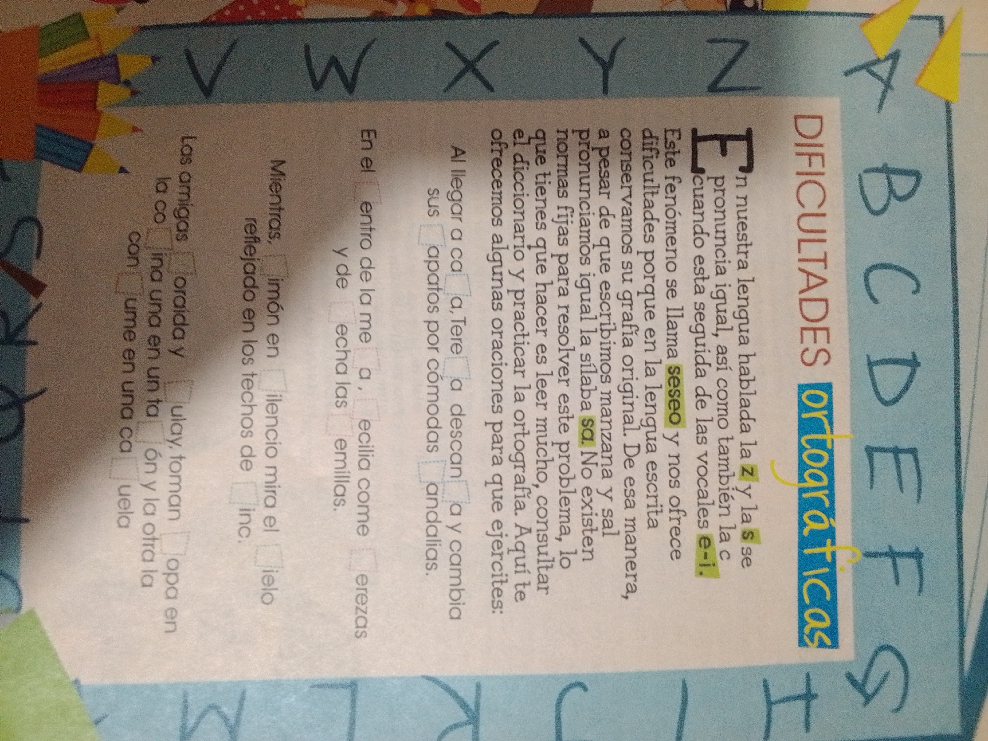 The image shows a page from a Spanish language learning book, likely for children. The page features a section titled "DIFICULTADES ORTOGRÁFICAS" (Orthographic Difficulties). The text discusses phonetic challenges in Spanish, specifically regarding the sounds of 's', 'z', 'y', and 'll'. There are also multiple-choice questions or fill-in-the-blank exercises presented as checkboxes next to sentence fragments. The page has a light yellow background with some graphic elements that appear to be colored pencils. The letters of the alphabet are also displayed on the right side of the page in large, stylized lettering. There are no people or specific locations visible in the image. The image does not contain any information related to La Tagua, Venezuela.