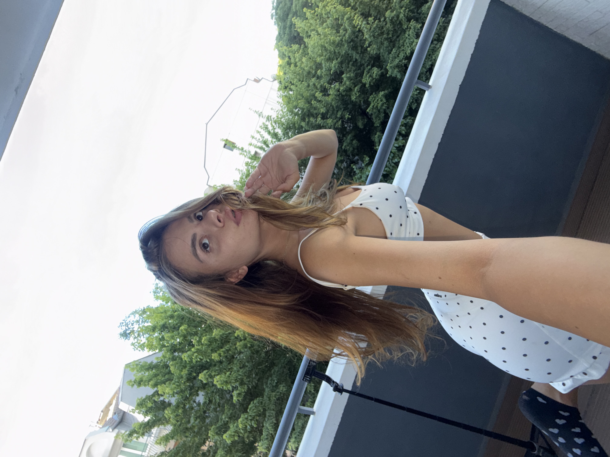 A young woman with long, auburn hair poses for a selfie on a balcony. She is wearing a white polka dot crop top and matching shorts. Her right hand is raised to her face, and she is looking at the camera with a playful expression. In the background, lush green trees and buildings are visible. The balcony has a grey railing. The setting appears to be a residential area, possibly an apartment building, in the city of Plovdiv, Bulgaria.
